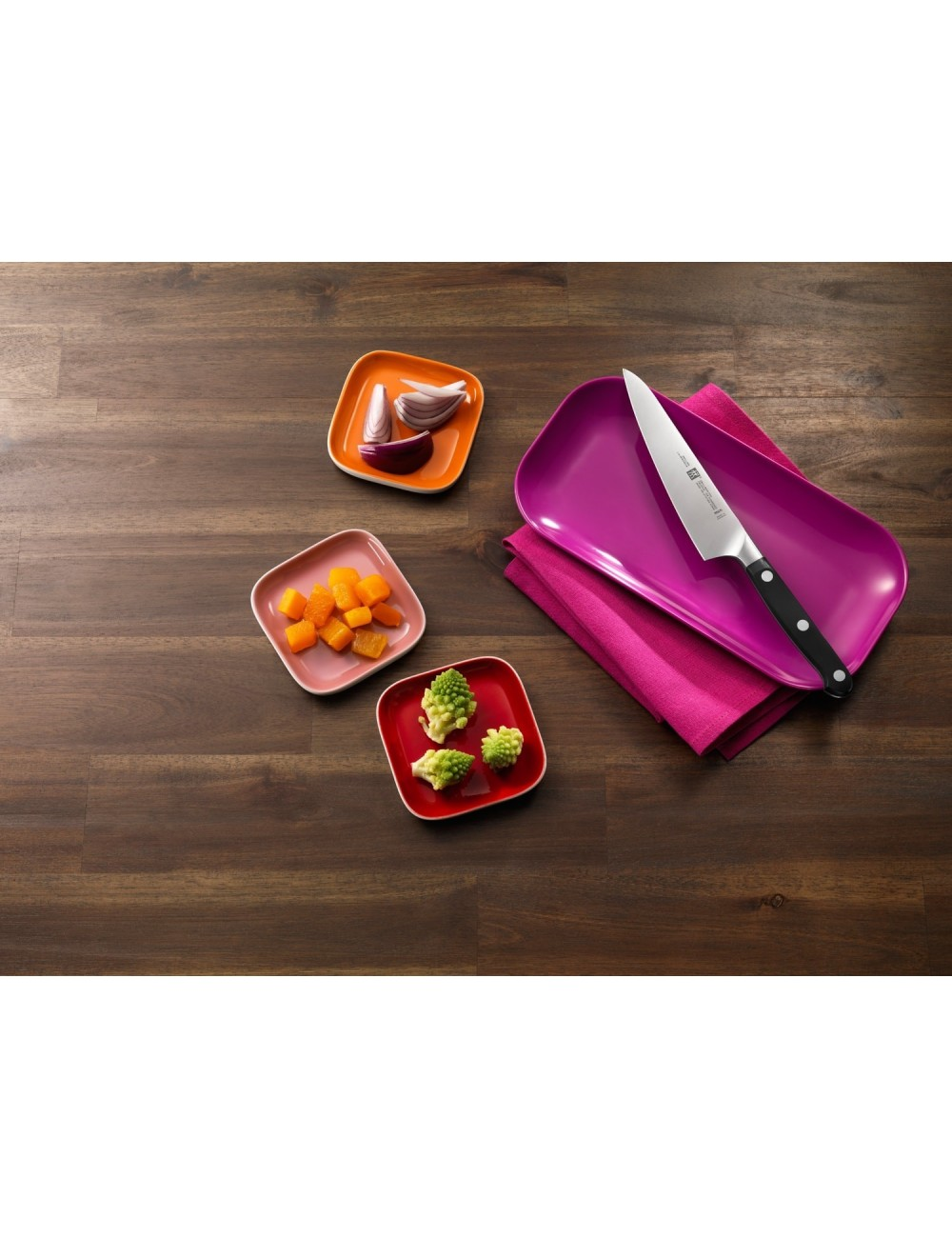 Drewniany stół z talerzami składników: cebula, dynia, romanesco, nóż na różowej serwetce i fioletowym talerzu.