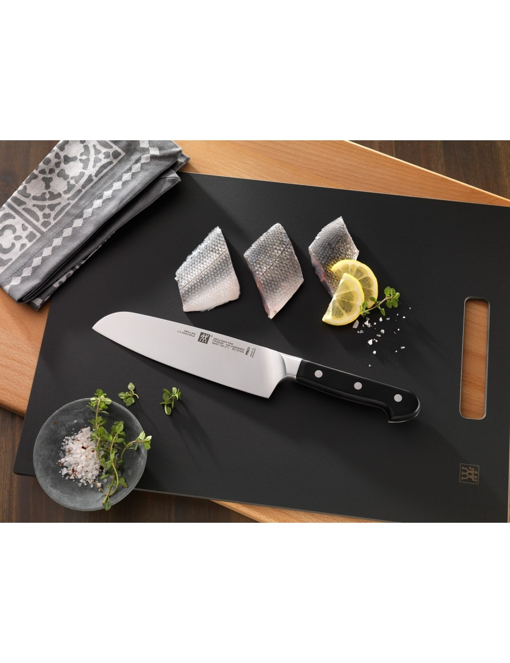Nóż na czarnej desce do krojenia z filetami rybnymi, plasterkami cytryny, ziołami i solą. Szara, wzorzysta ściereczka obok.