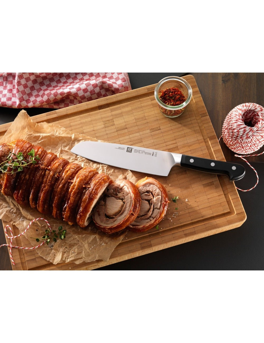 Krojona wieprzowina pieczona na drewnianej desce z nożem, ziołami, przyprawami i sznurkiem. Czerwono-biała krata w tle.