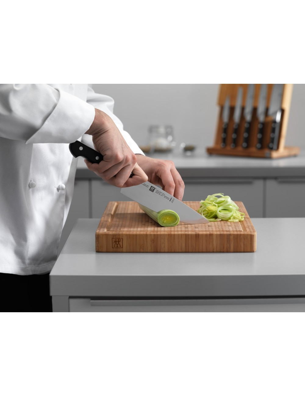 Szef kuchni w białym uniformie kroi por na drewnianej desce do krojenia dużym nożem. Inne noże są w tle.