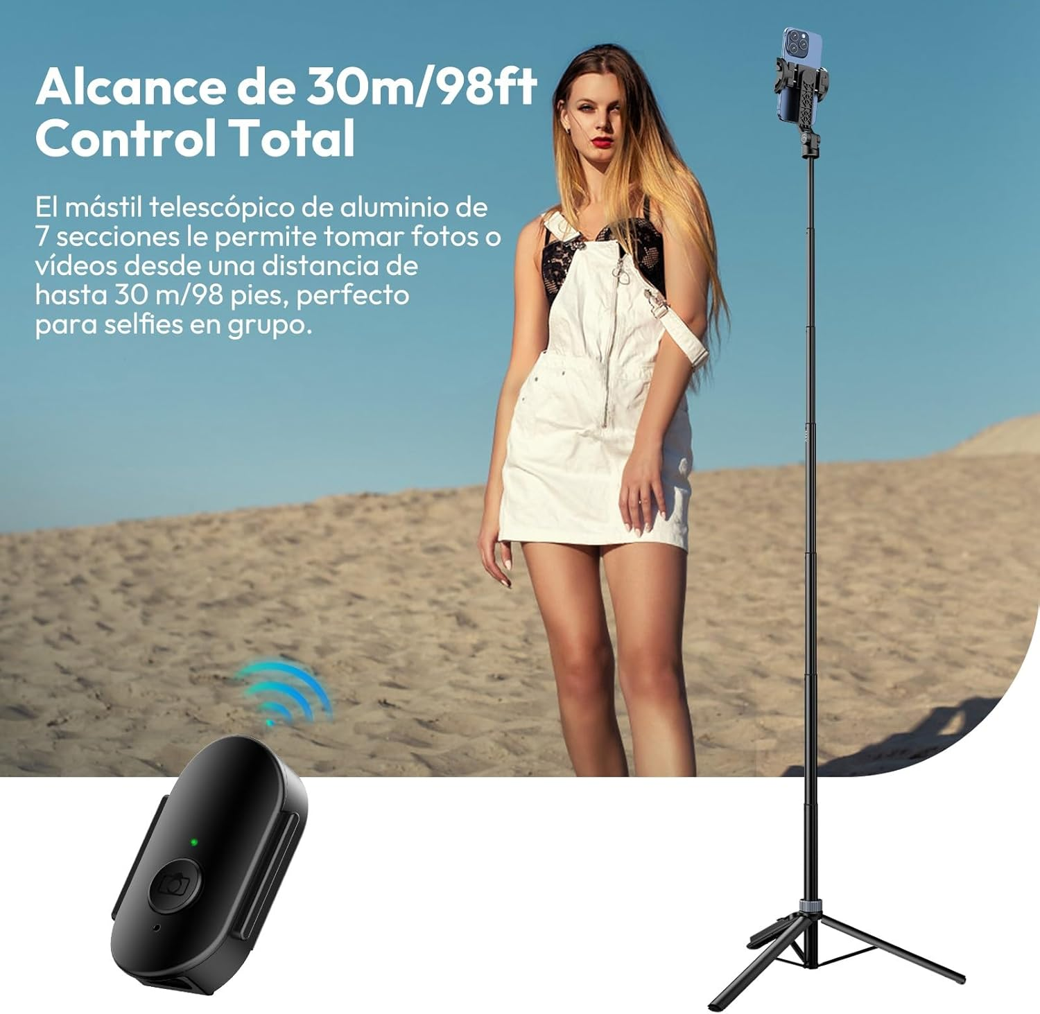 Una mujer está de pie con un palo para selfies. Un control remoto está en primer plano. El fondo es una playa arenosa bajo un cielo azul.
