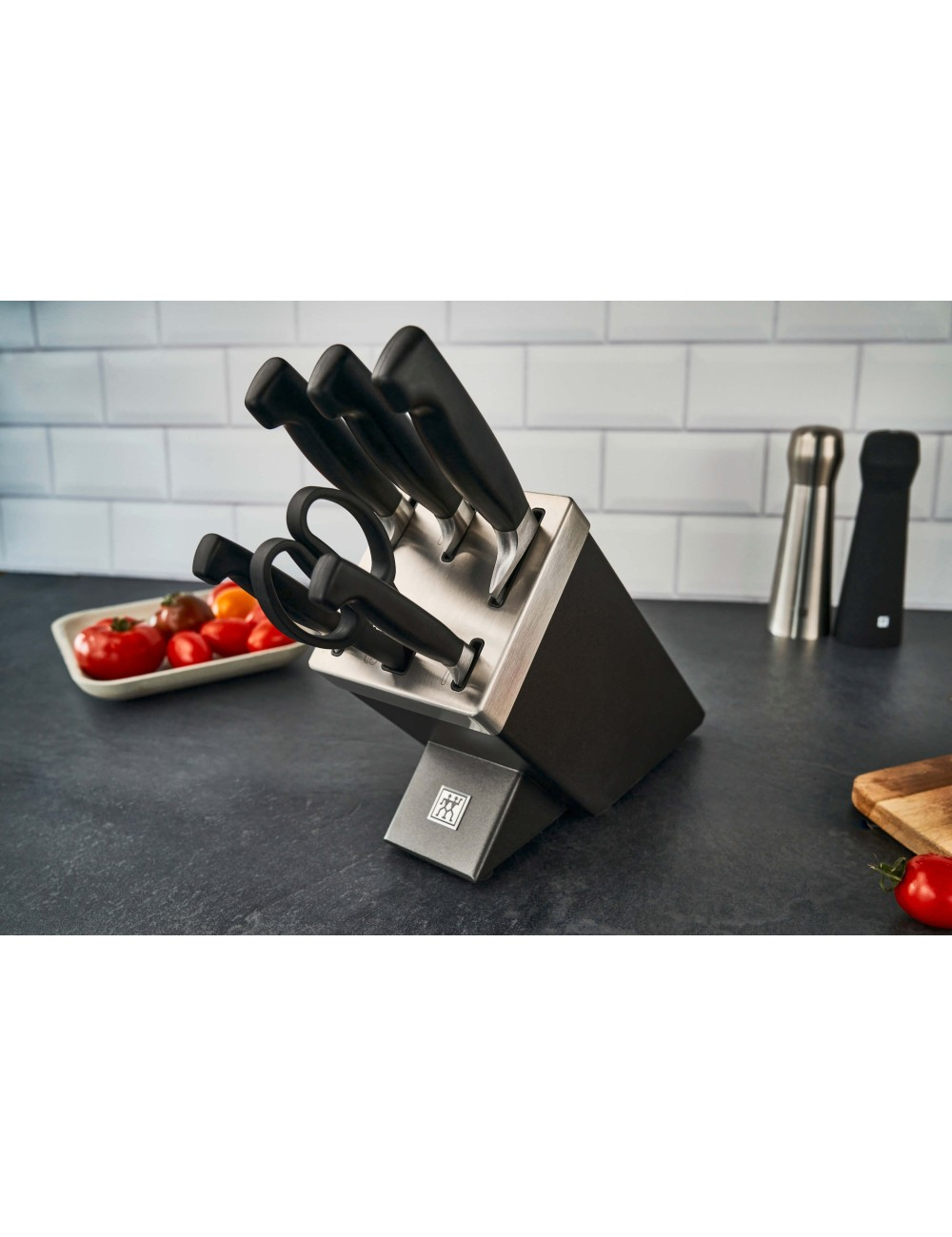 Blok noży z nożami i nożyczkami na ciemnym blacie. Obecne są również pomidory oraz solniczki i pieprzniczki.