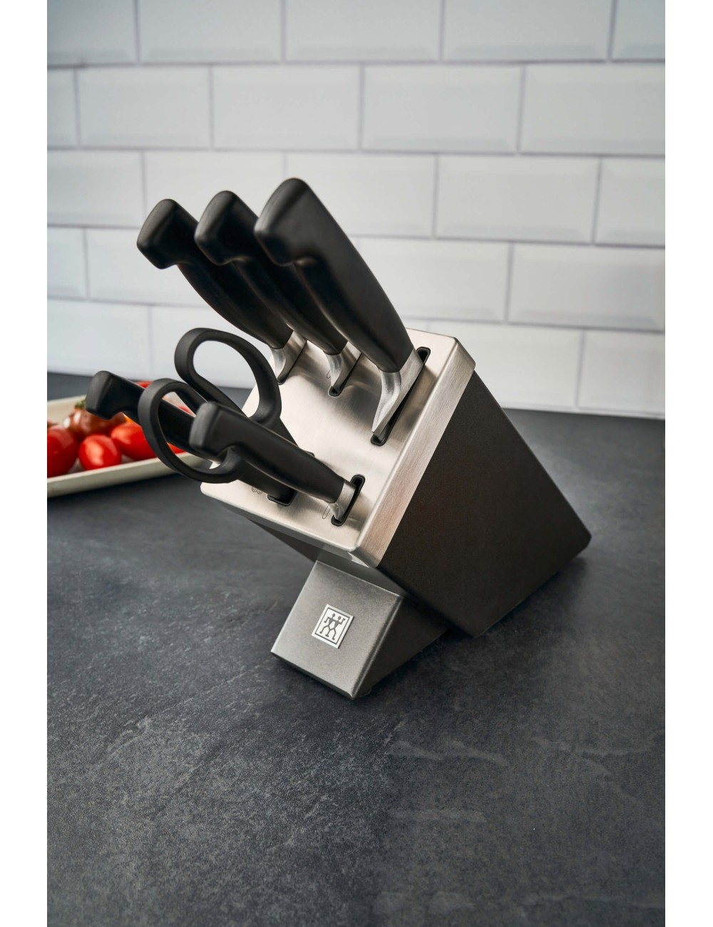 Blok noży z nożami i nożyczkami na ciemnym blacie. Talerz pomidorów w tle. Ściana z białych płytek.