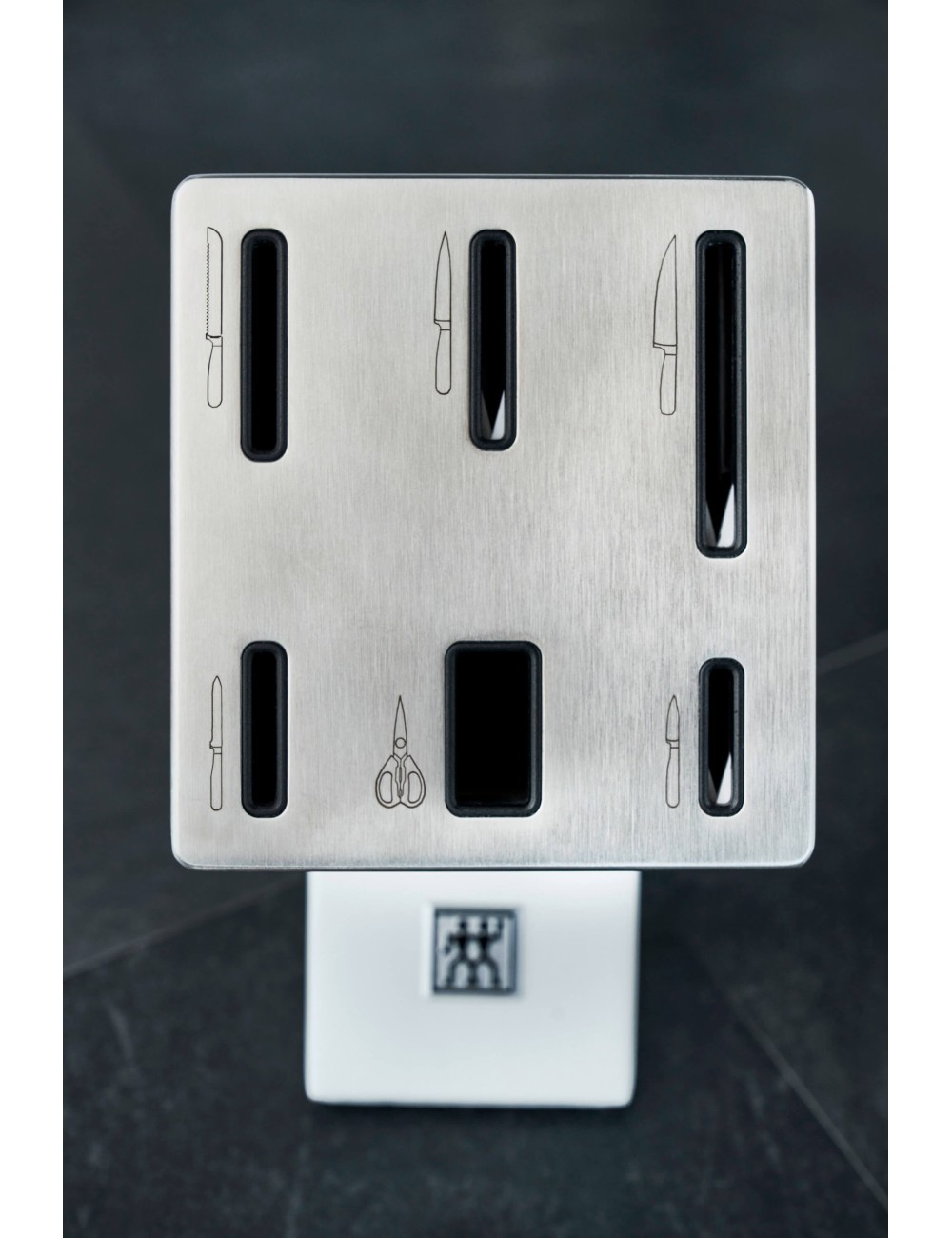 Blok na noże ze stali nierdzewnej z otworami, mieszczący noże i nożyczki. Biała podstawa z logo spoczywa poniżej na ciemnej powierzchni.