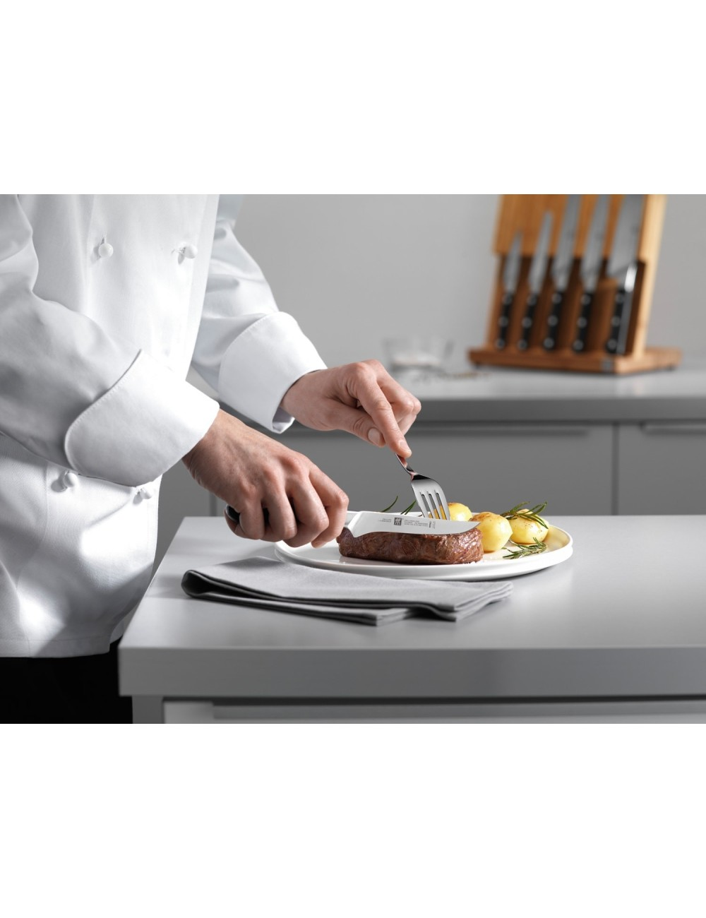 Szef kuchni kroi stek nożem i widelcem. Stek leży na talerzu z ziemniakami i rozmarynem. Noże są w tle.