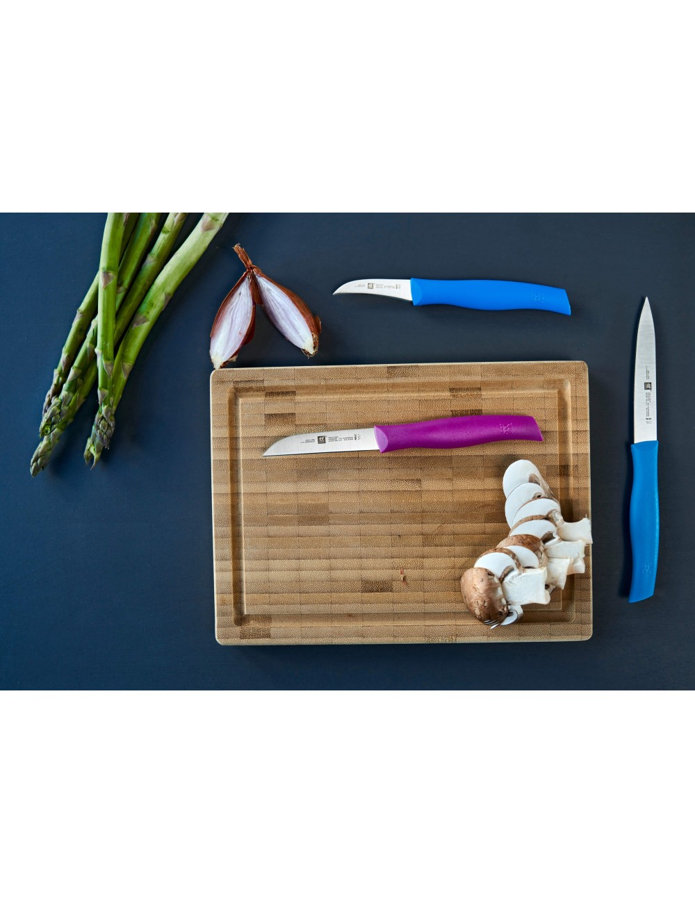 Deska do krojenia z nożami, szparagami, szalotką i pokrojonymi pieczarkami. Noże mają niebieskie i fioletowe uchwyty.