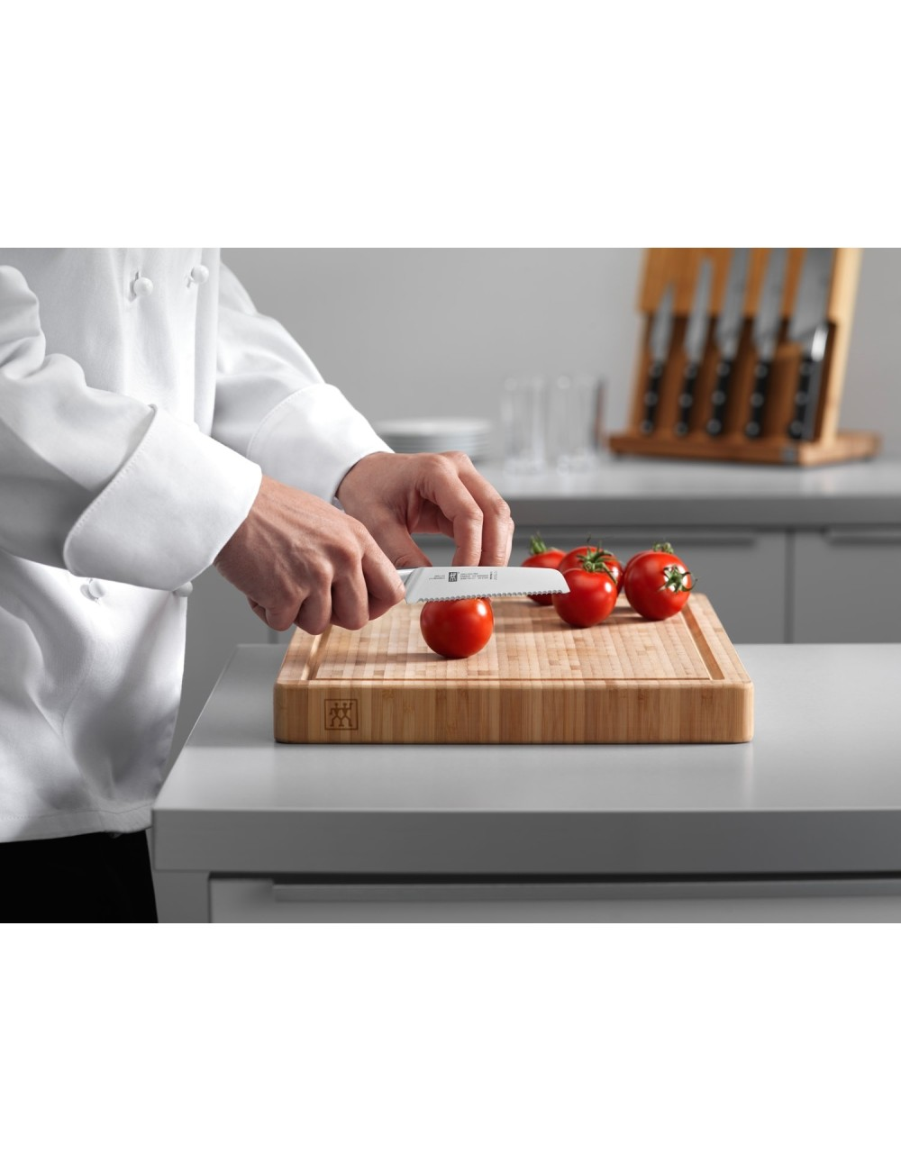 Szef kuchni kroi pomidora nożem z ząbkowanym ostrzem na drewnianej desce do krojenia. Widoczne są inne pomidory. W tle blok z nożami.