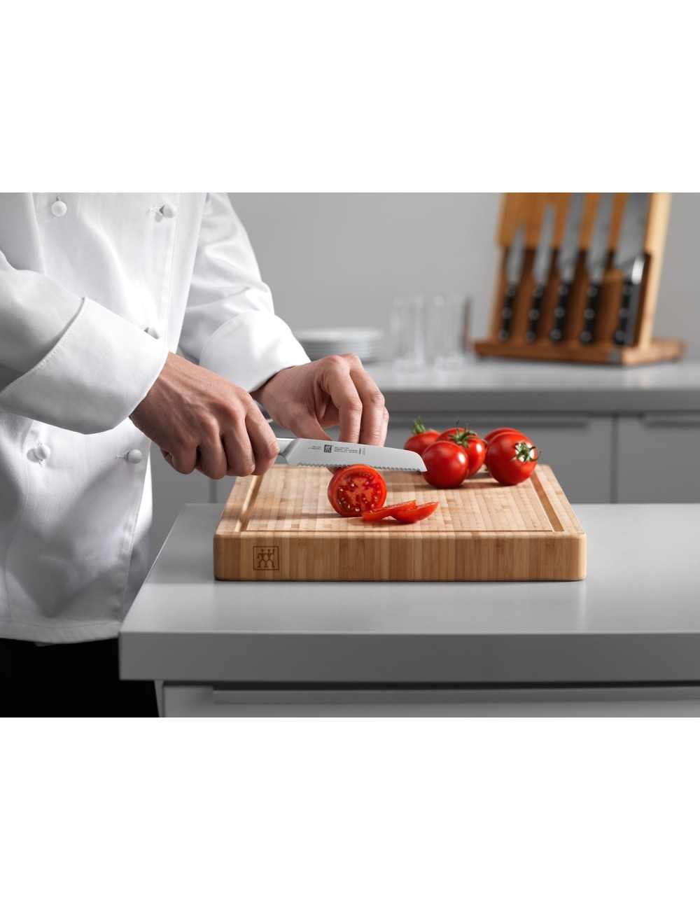 Szef kuchni kroi pomidora na drewnianej desce do krojenia. Inne pomidory i noże są w tle. Szef kuchni ma na sobie biały fartuch.