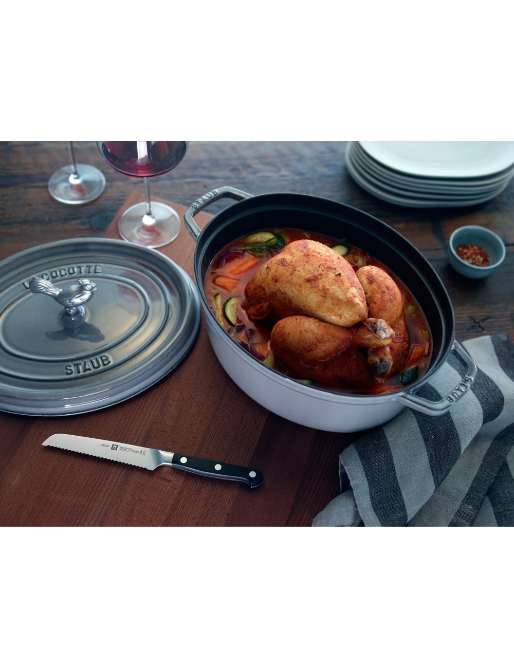 Ugotowany kurczak w garnku Staub z warzywami. Obecne są również nóż, kieliszki do wina, talerze i miska z przyprawami.