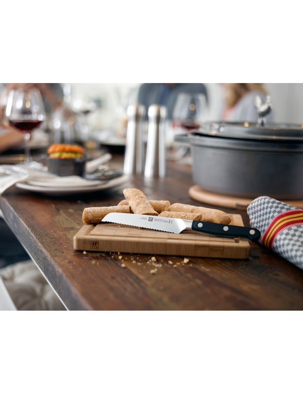 Nakrycie stołu z paluszkami, nożem do chleba na drewnianej desce i garnkiem z pokrywką. Widoczne są inne potrawy i kieliszki do wina.