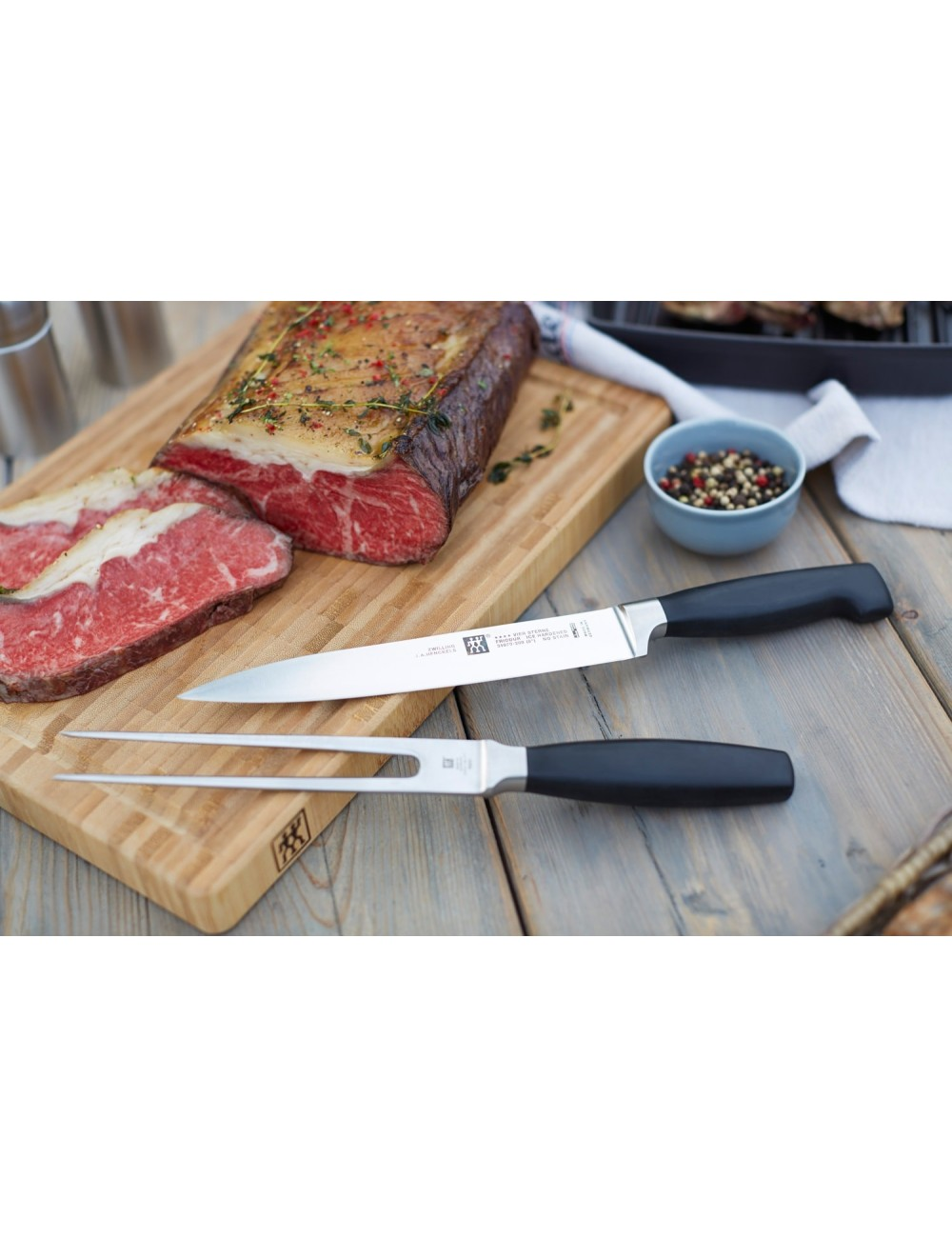 Deska do krojenia z gotowanym i pokrojonym mięsem. Nóż i widelec leżą na drewnianym stole. Miska z ziarnami pieprzu jest na stole.