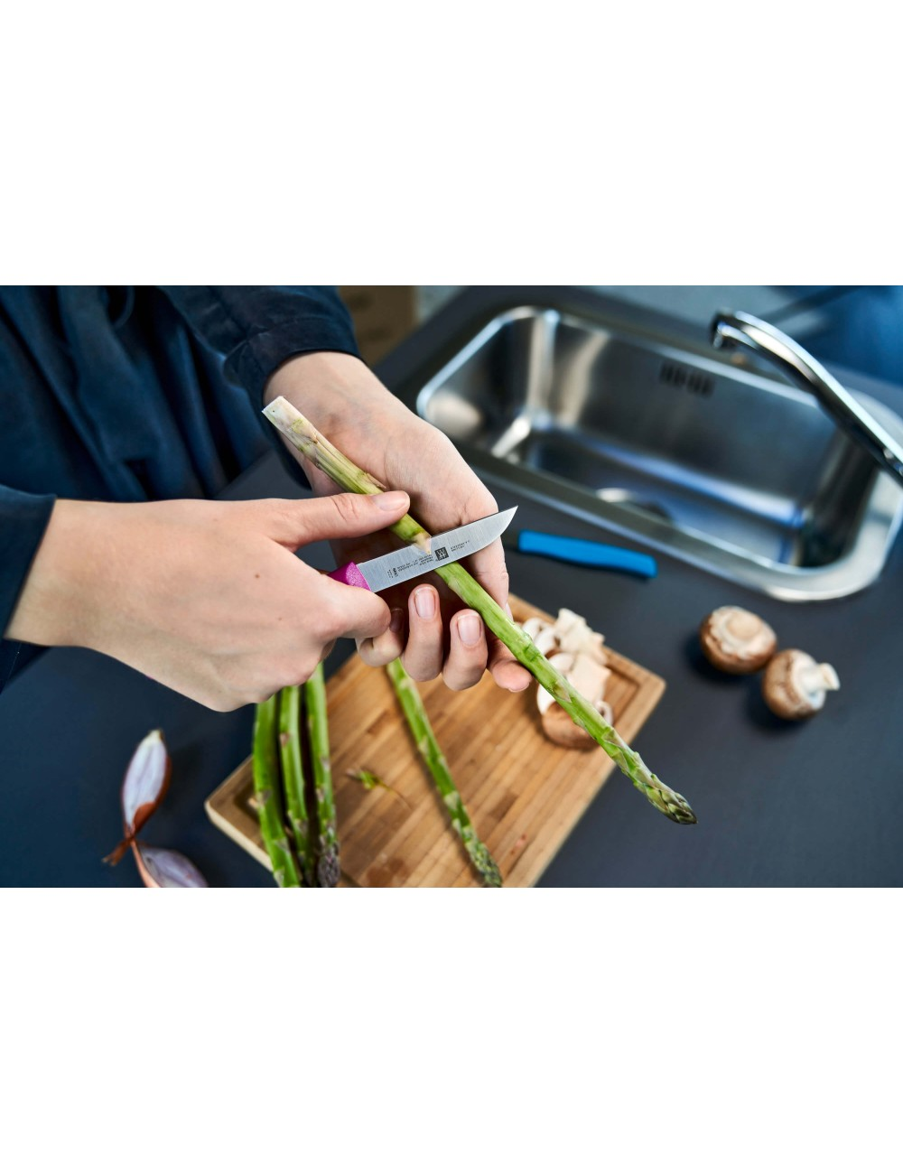 Ręce obierają szparagi nożem. Posiekane grzyby i zlew znajdują się w tle. Scena rozgrywa się na ciemnym blacie.