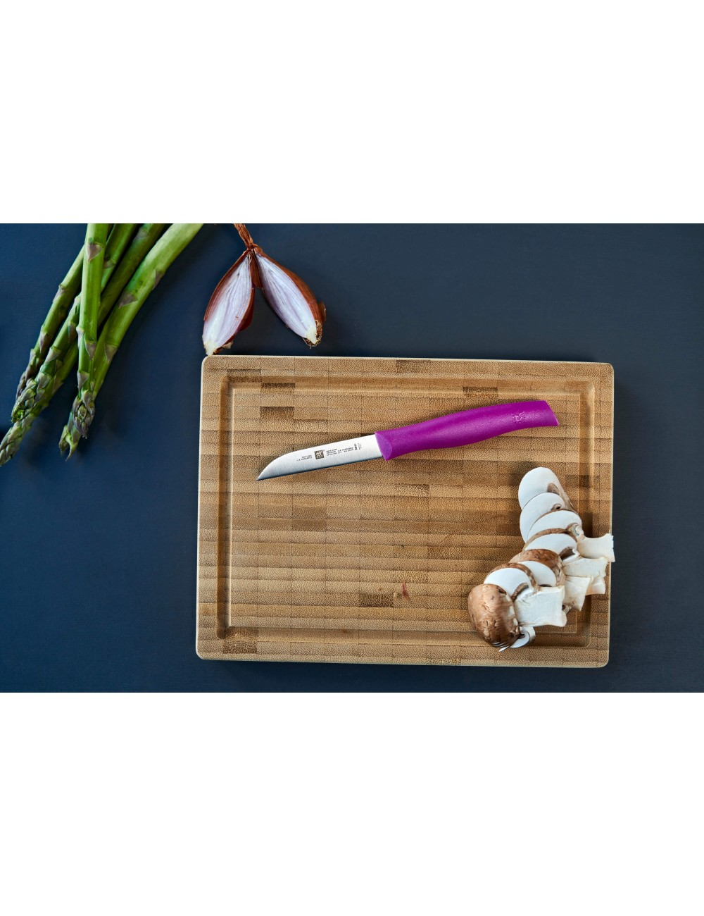 Deska do krojenia z nożem, pokrojone pieczarki, szparagi i cebula na ciemnoniebieskiej powierzchni.