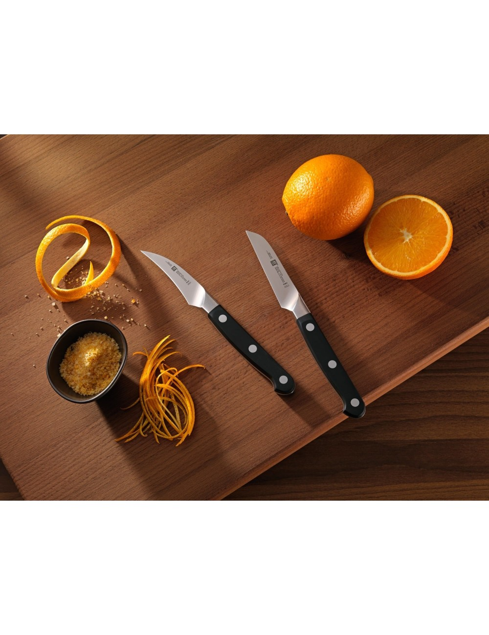 Dwa noże, pomarańcze (całe i przekrojone), skórka pomarańczowa i miska z cukrem pomarańczowym na drewnie.