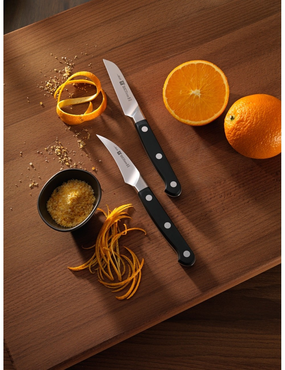 Dwa noże, skórka pomarańczowa, pomarańcze pokrojone i całe oraz miska ze skórką na drewnie.