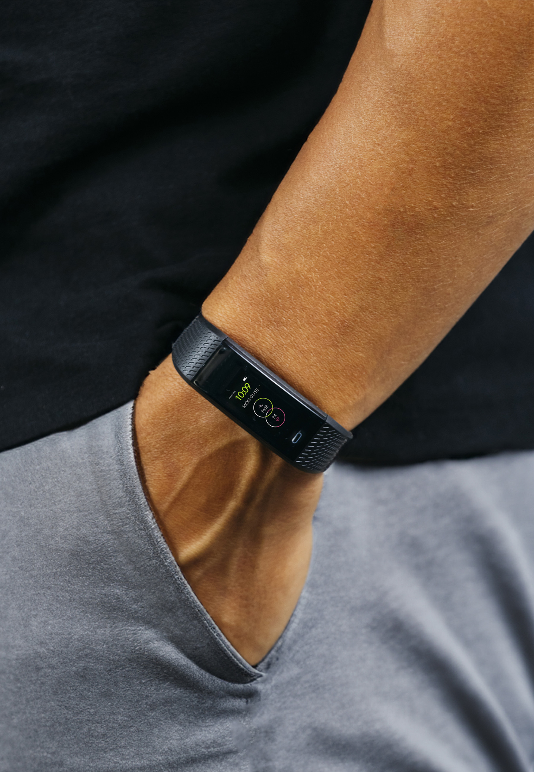 Ein Arm mit einem schwarzen Fitnesstracker. Die Uhrzeit ist 10:09 Uhr. Die Person trägt ein schwarzes Hemd und graue Hose.