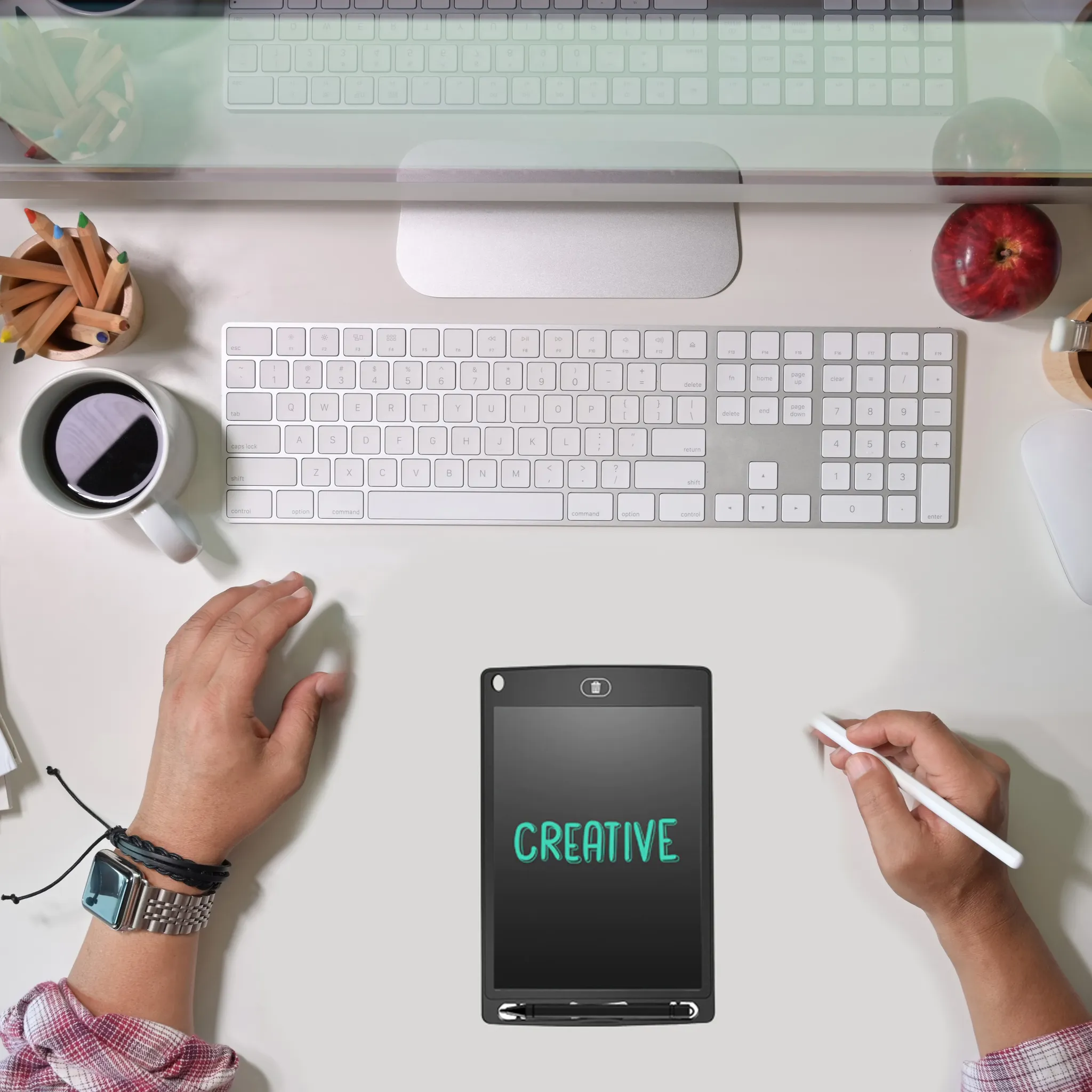 Schreibtisch mit Tastatur, Tablet mit CREATIVE, Hände, Kaffee, Stifte, Apfel. Hände halten Stift und Smartwatch.