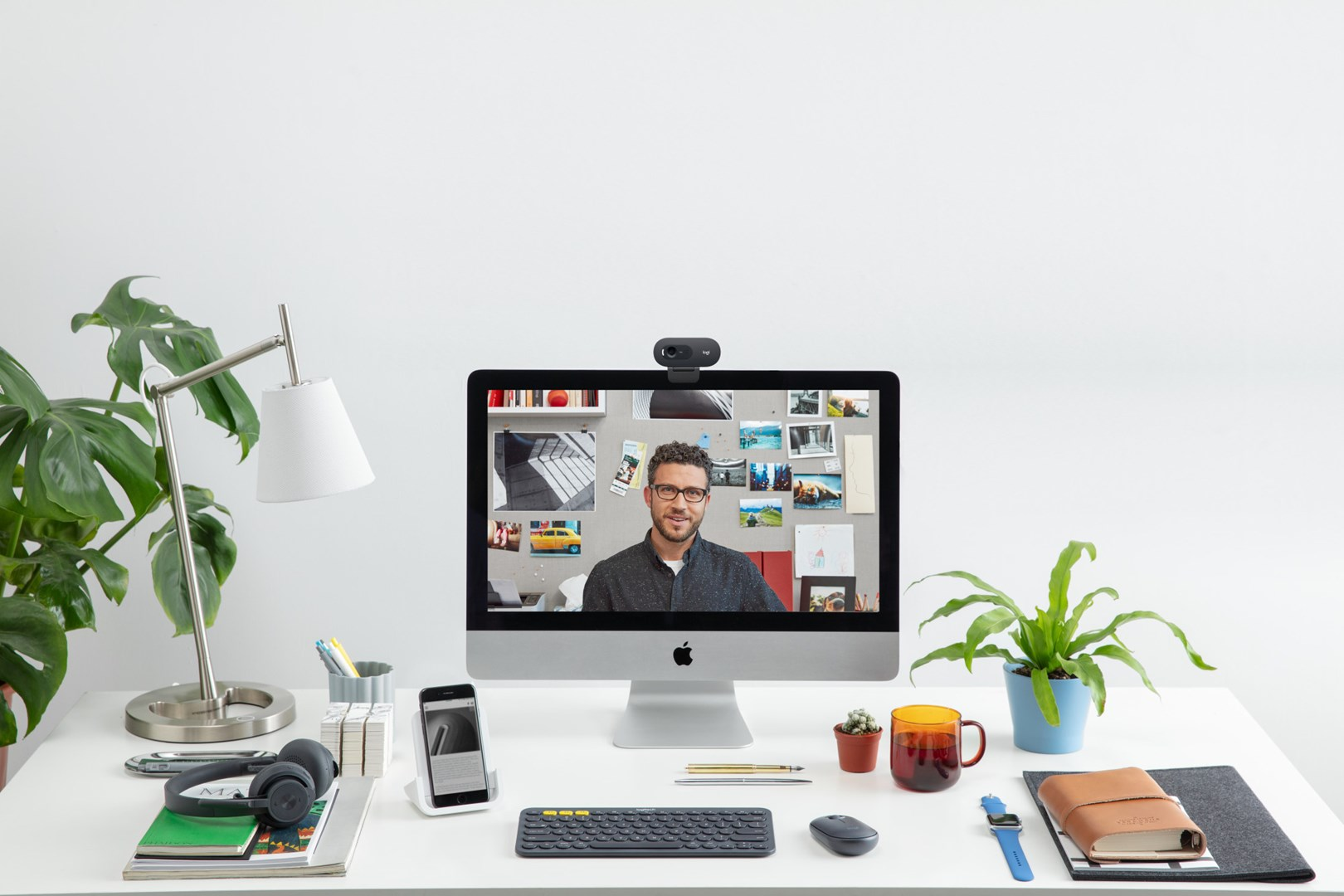 Mężczyzna na ekranie komputera siedzi przy biurku z roślinami, lampą i gadżetami technologicznymi.