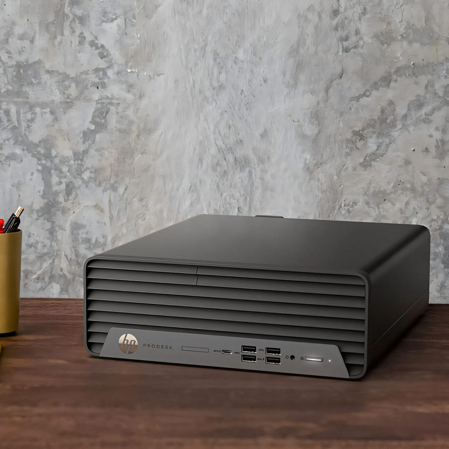 Una computadora HP ProDesk negra sobre un escritorio de madera marrón. Una pared de hormigón al fondo. Un portalápices a la izquierda.