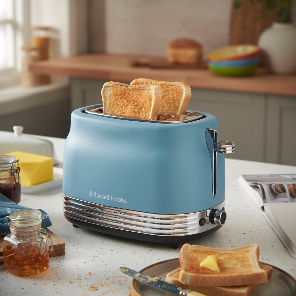 Ein blauer Toaster mit Toastbrot steht auf einer Theke mit Butter, Marmelade und Toast. Küchenszene.