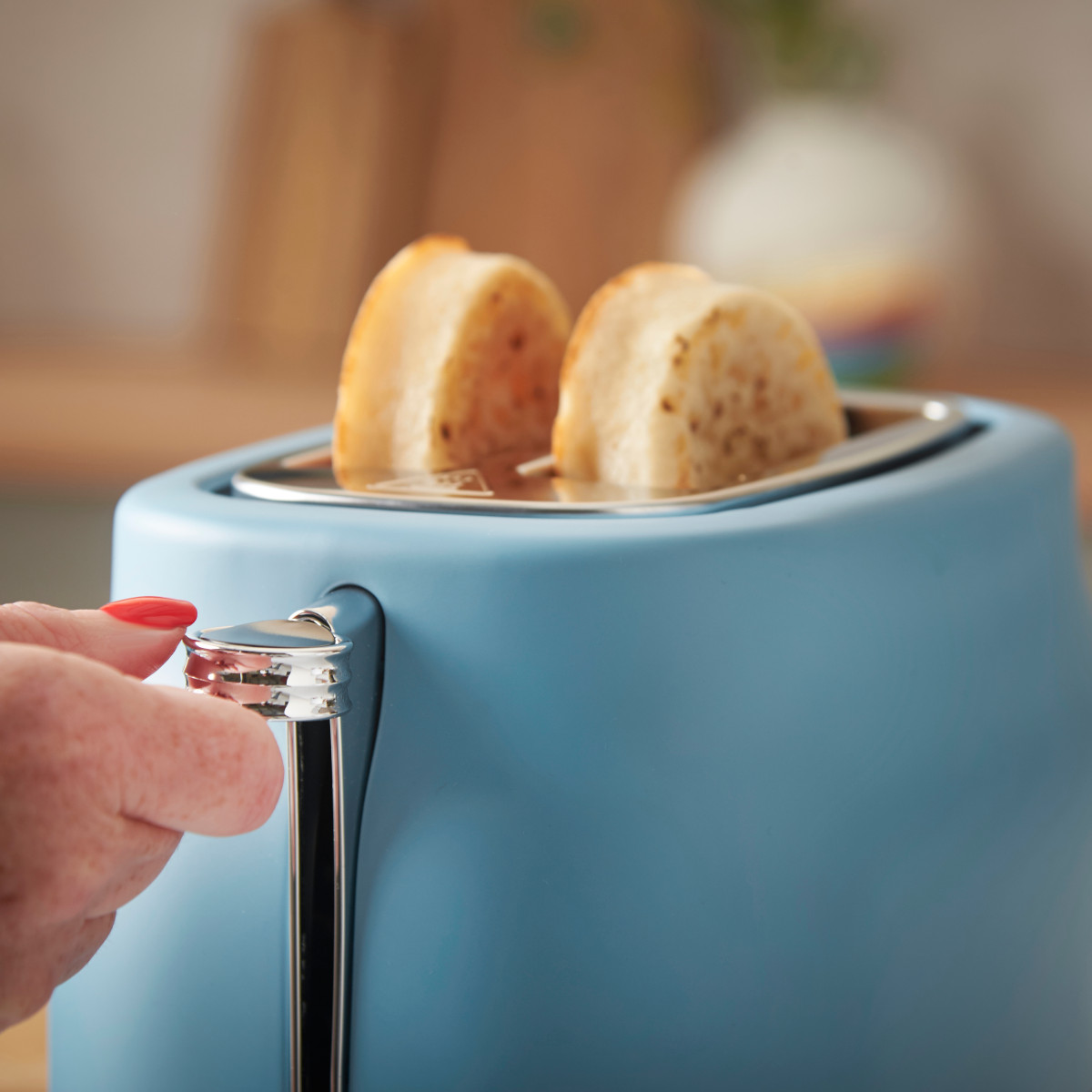 Eine Hand mit rotem Nagellack drückt einen Hebel an einem blauen Toaster mit zwei Toastscheiben.