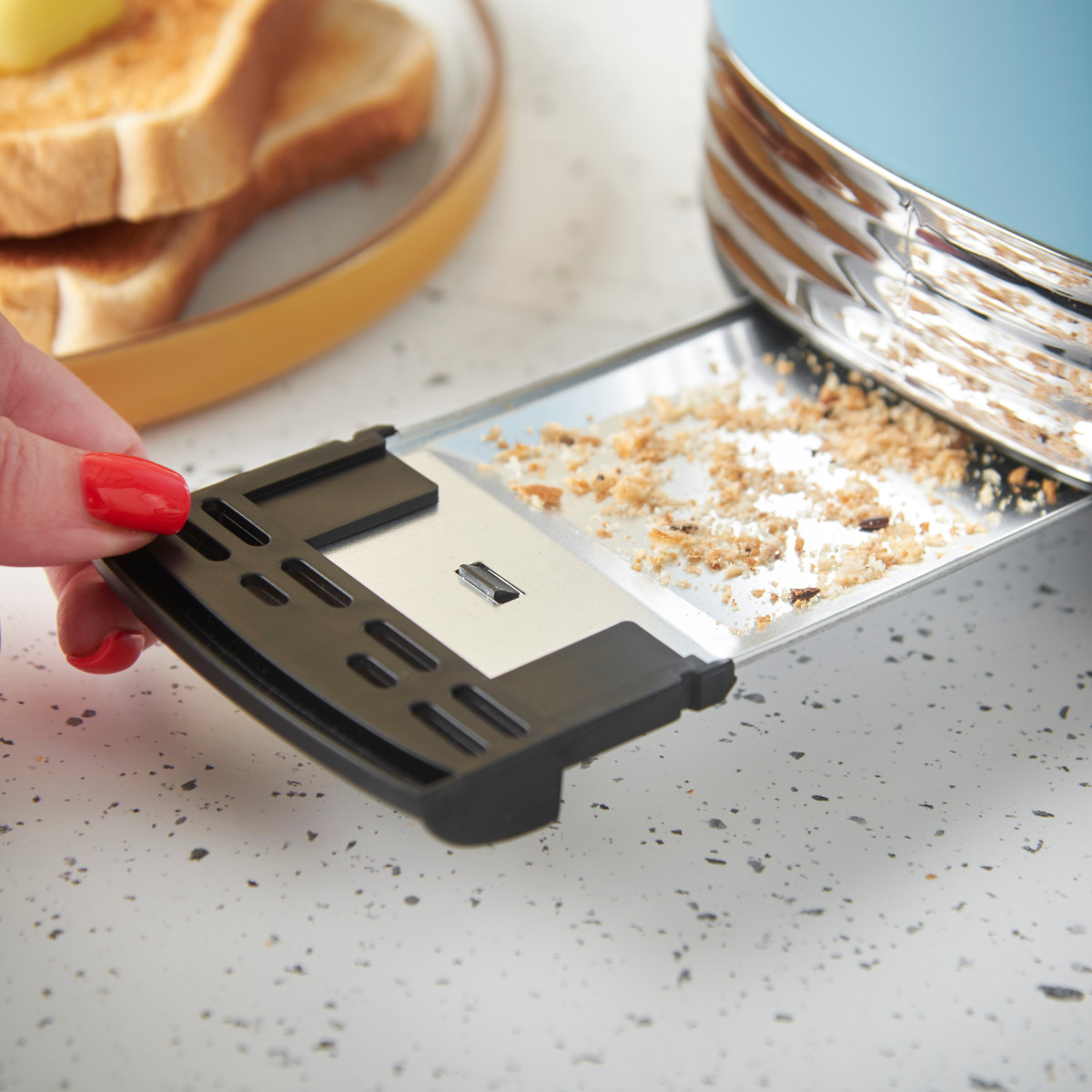 Eine Hand mit roten Nägeln zieht eine Krümelschublade aus einem blauen Toaster. Brot und Butter liegen auf einem Teller im Hintergrund.