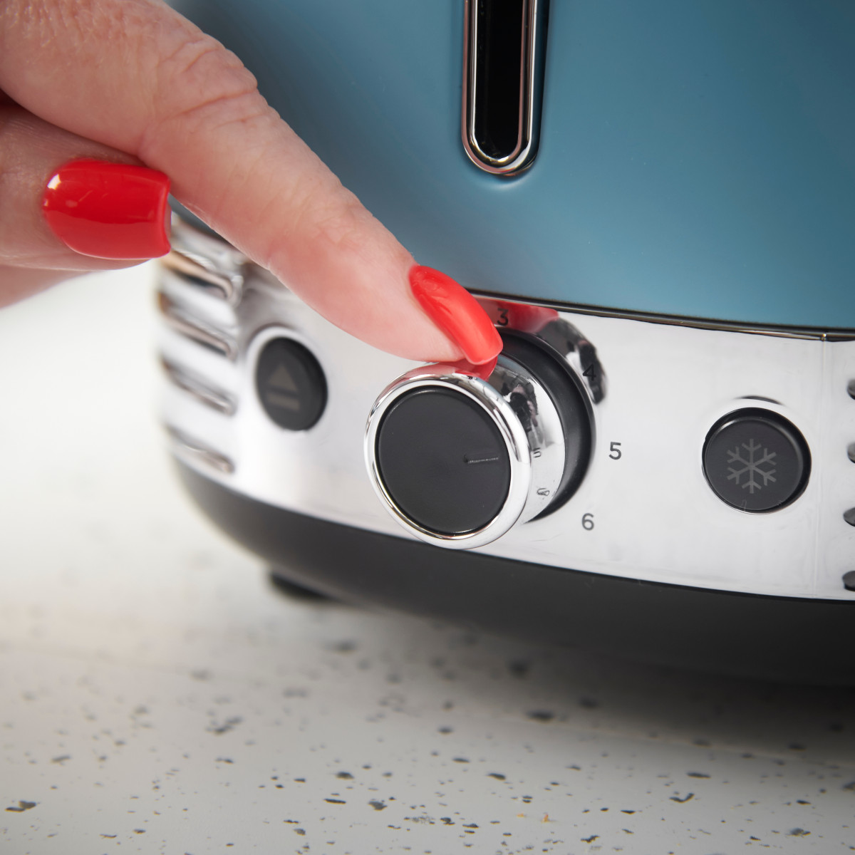 Eine Hand mit roten Nägeln dreht einen Toaster-Regler. Der Toaster ist blaugrün und verchromt. Die Zahlen 5 und 6 sowie ein Schneeflockensymbol sind sichtbar.