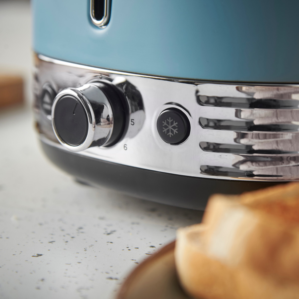 Nahaufnahme eines türkisfarbenen Toasters mit Chromakzenten. Er hat einen Knopf, Knöpfe und Schlitze für Brot. Toastscheiben befinden sich im Vordergrund.