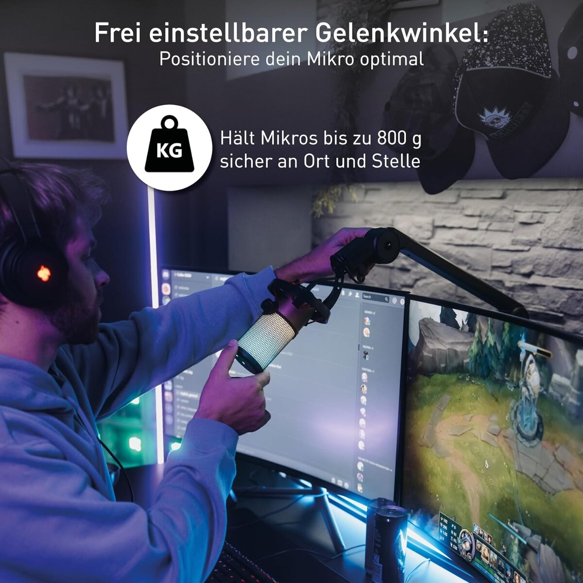 Ein Mann justiert ein Mikrofon an einem Ständer vor zwei Computerbildschirmen, von denen einer ein Spiel zeigt. Im Hintergrund sind Hüte zu sehen.