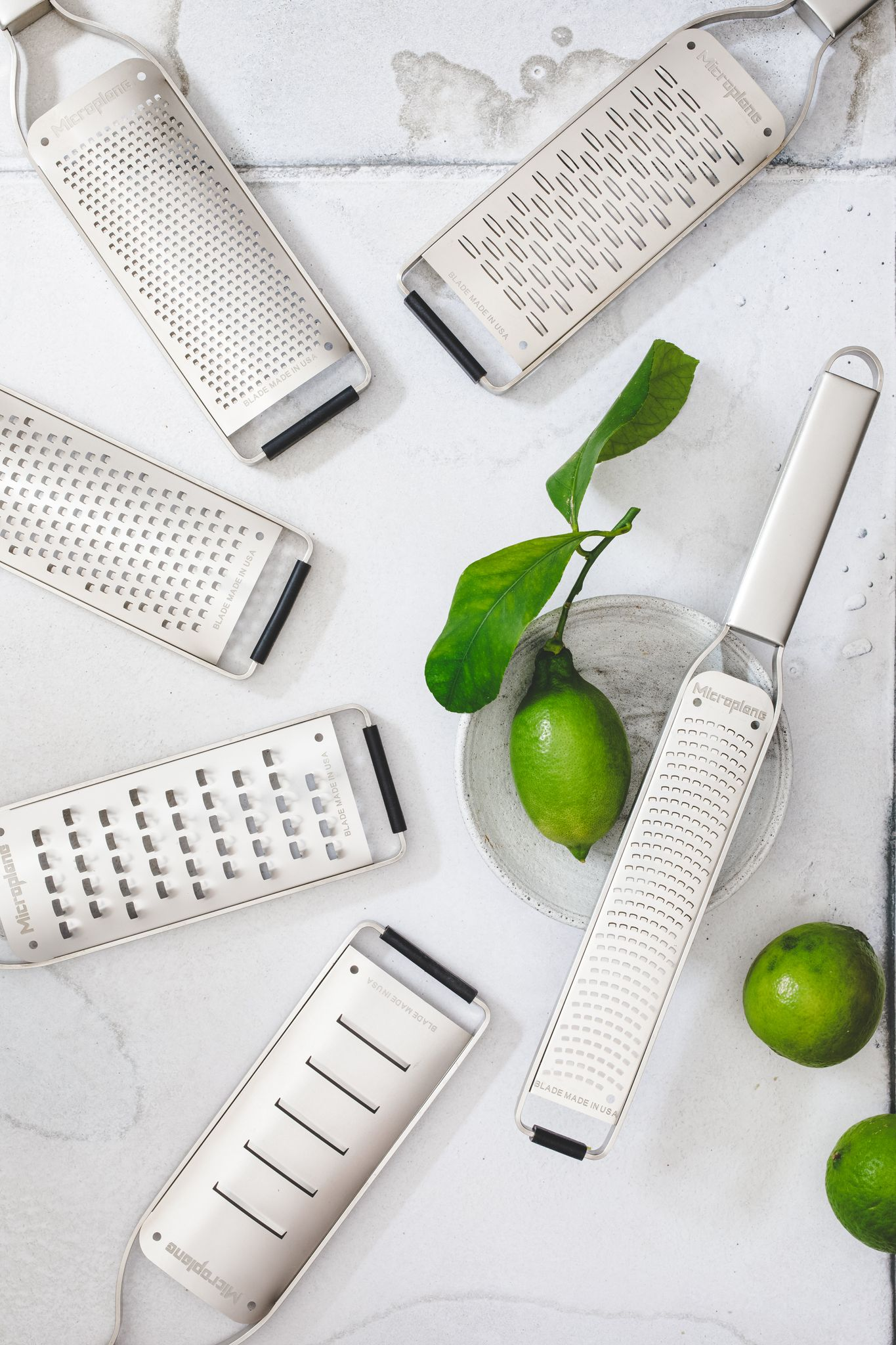 Mehrere Microplane-Reiben sind um eine Limette, eine Schüssel und Blätter auf einer weißen Oberfläche verstreut. Die Reiben sind silberfarben.