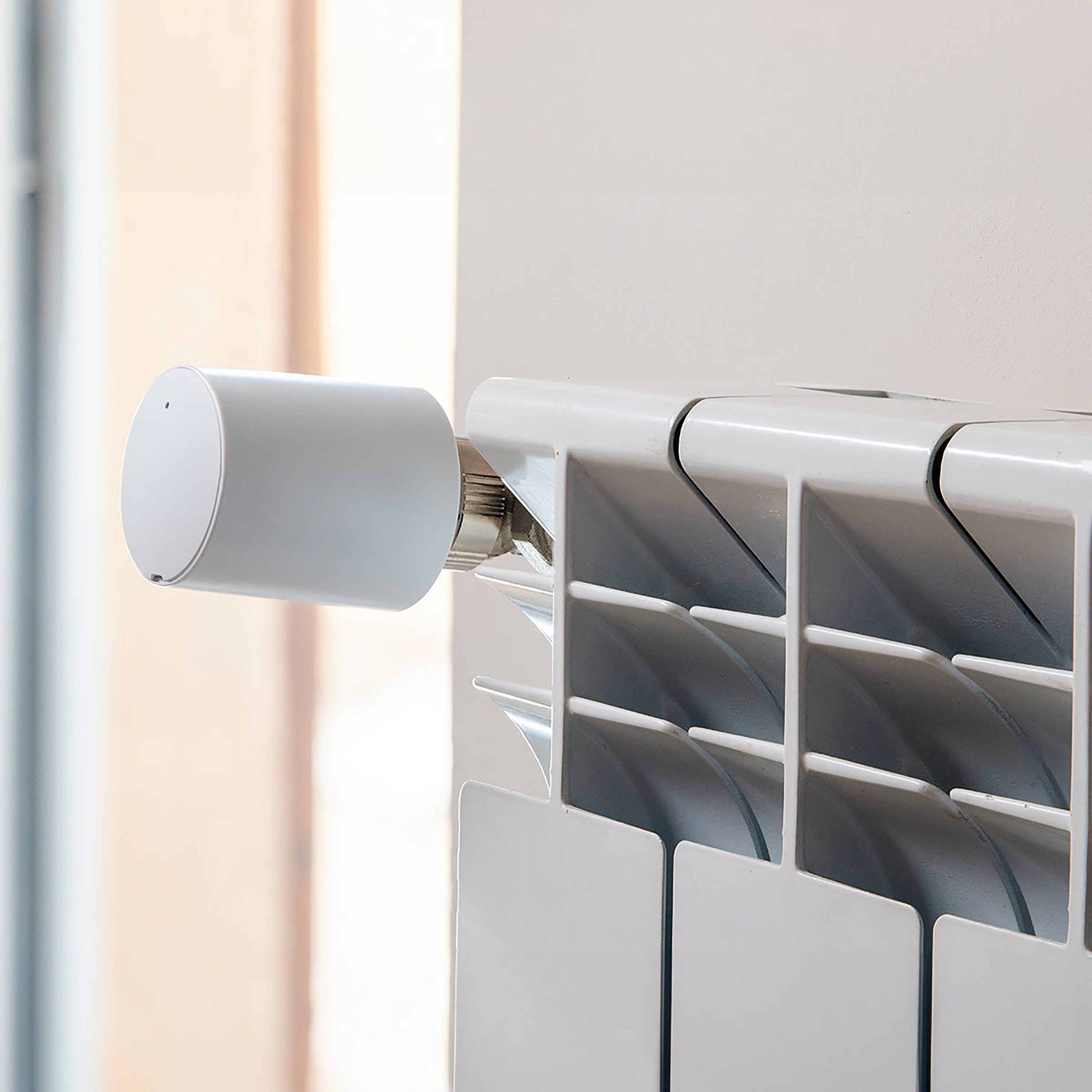 Biały grzejnik z białym, cylindrycznym termostatem. Tło to rozmyta ściana i okno.