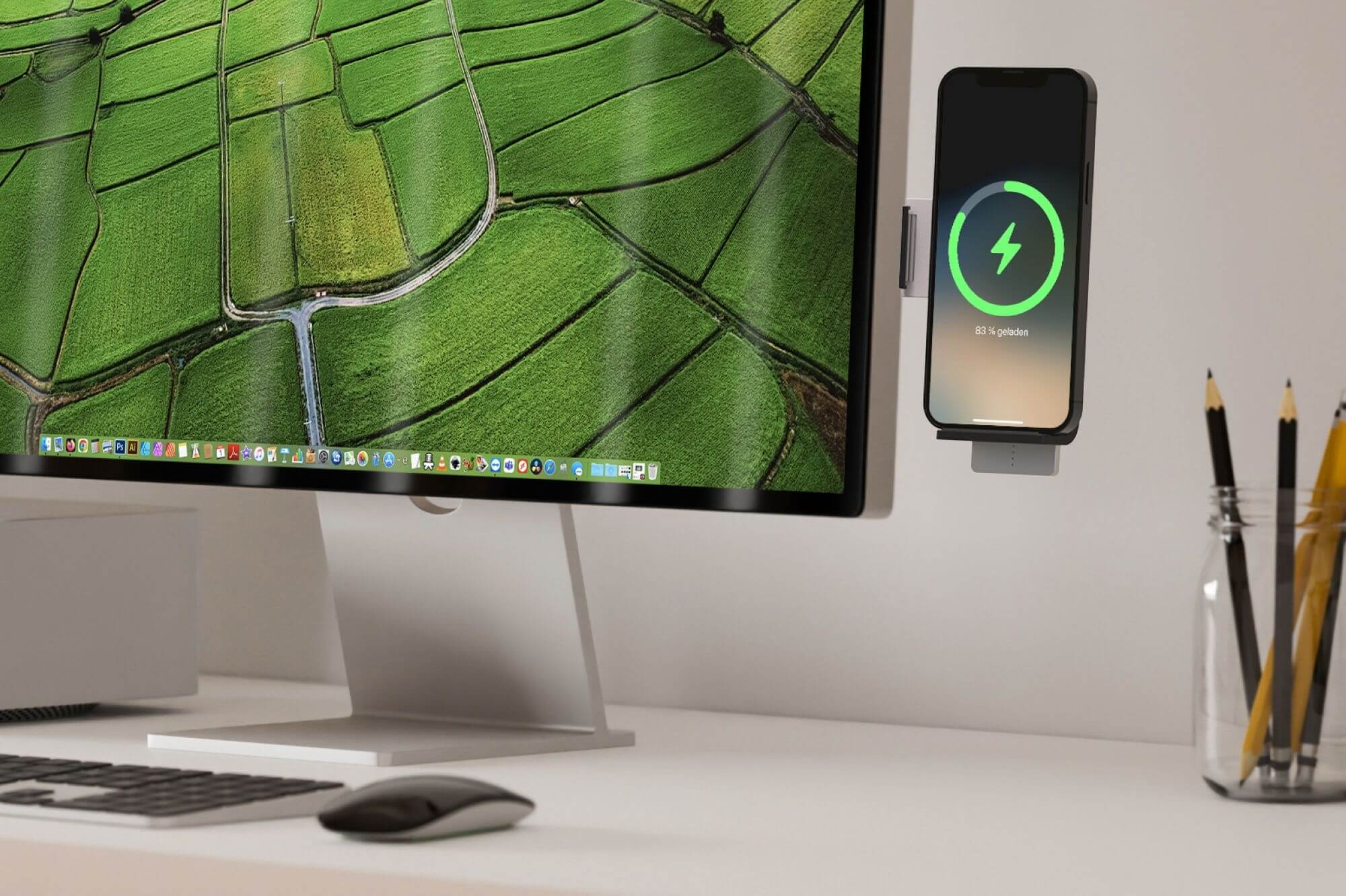 Ein Computermonitor zeigt ein grünes Feld. Ein Telefon wird geladen. Stifte in einem Glas stehen rechts. Eine Tastatur und Maus sind im Vordergrund.