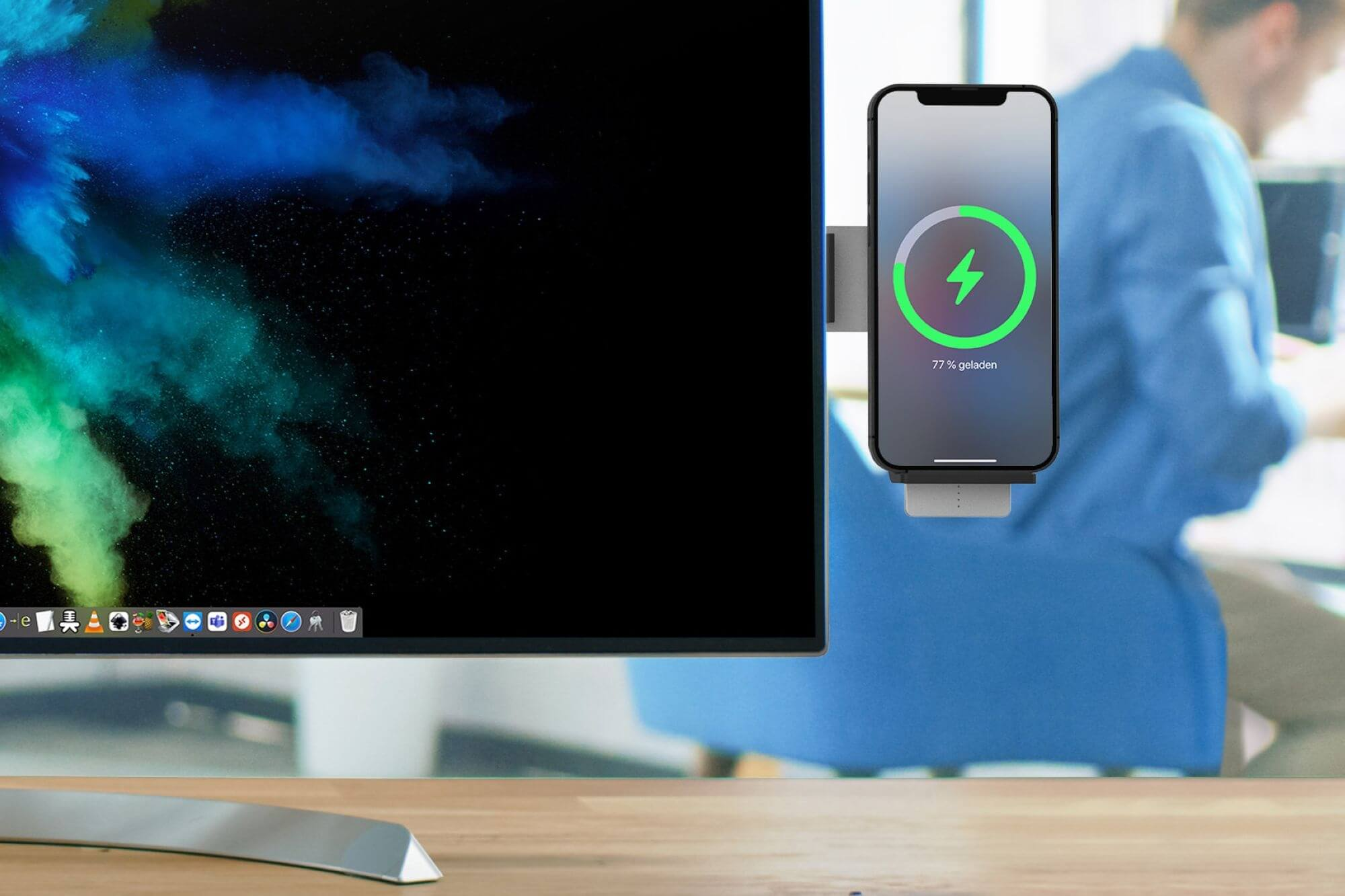 Ein Telefon, das auf einem Schreibtisch aufgeladen wird. Ein Monitor zeigt einen farbenfrohen Hintergrund. Eine Person in blauem Hemd im Hintergrund.