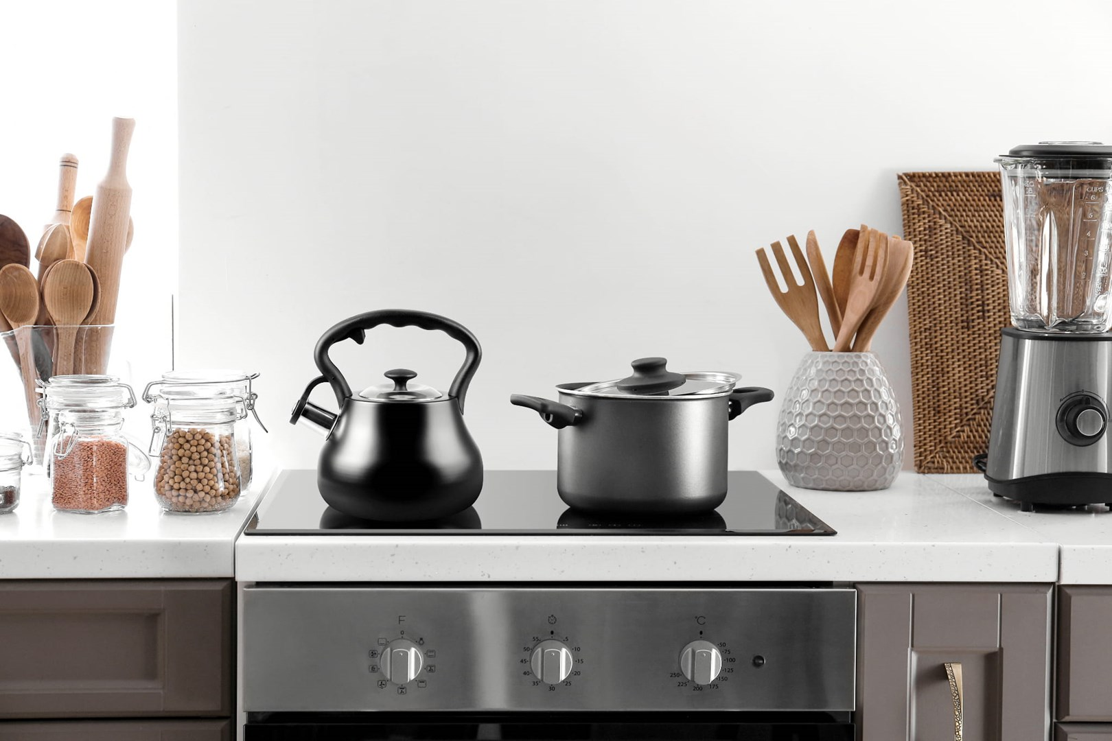 Scena kuchenna z czarnym czajnikiem, garnkiem, słoikami, blenderem i drewnianymi przyborami. Wszystko na białym blacie.
