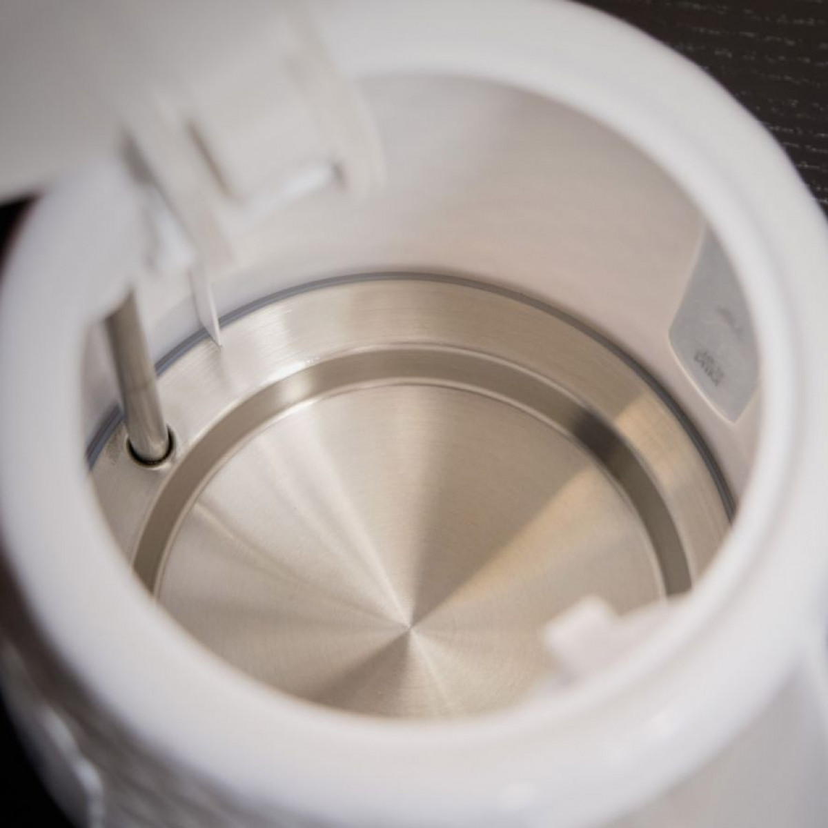 Widok wewnątrz białego czajnika elektrycznego. Wnętrze ze stali nierdzewnej, pokrywa otwarta. Widoczna metalowa rurka.