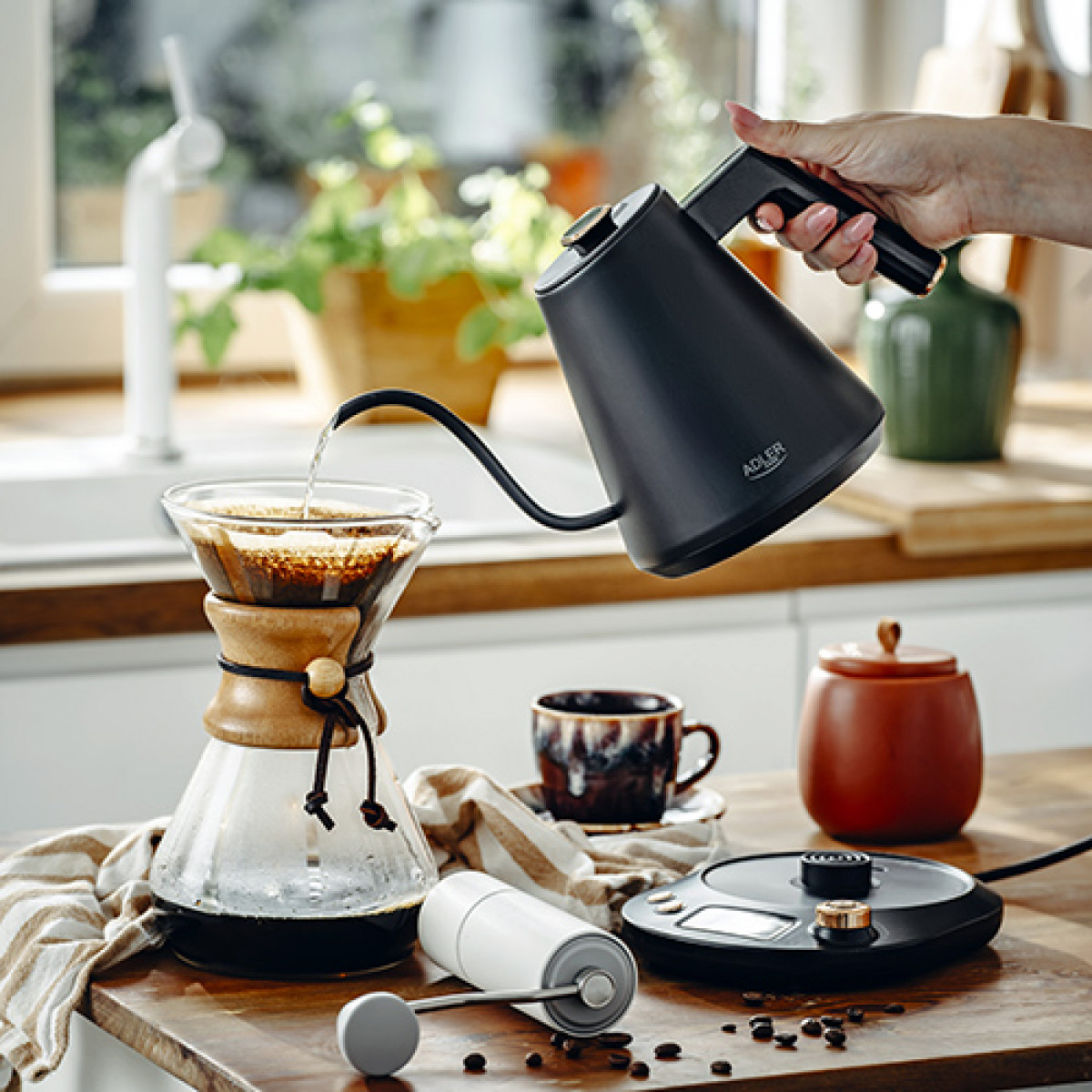 Ręka wlewa wodę z czarnego czajnika do ekspresu do kawy. Filiżanka, młynek i waga stoją na drewnianej powierzchni.