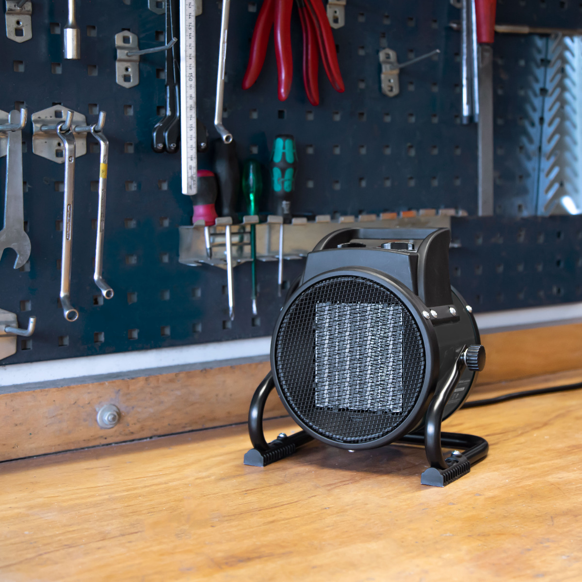 A black space heater sits on a wooden workbench. Tools hang on a pegboard in the background. Red pliers are visible.