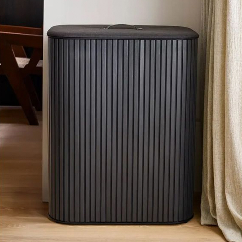 Una cesta de ropa oscura y gris con tapa y asa. Una silla de madera y una cortina beige están en el fondo.