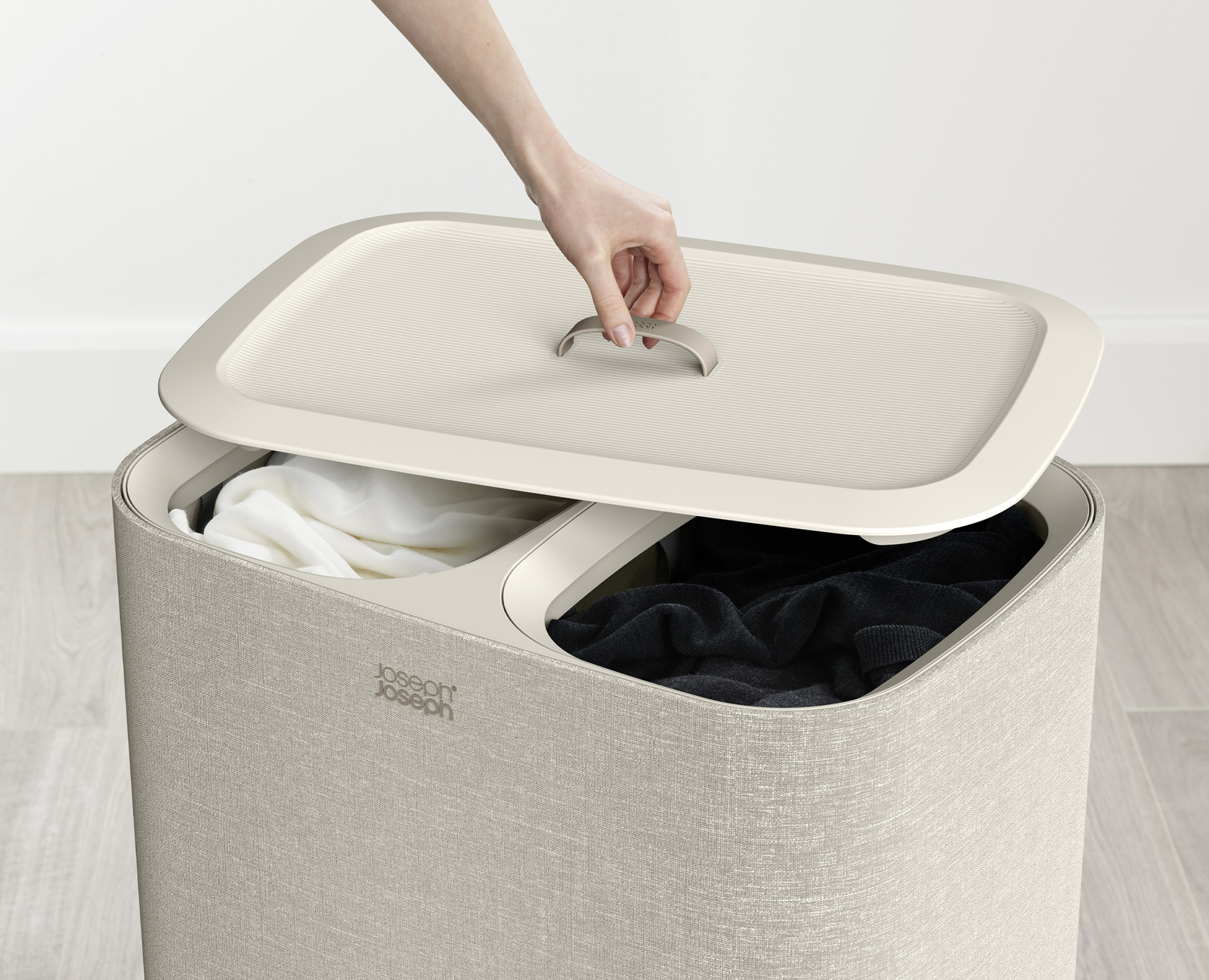 Una mano levanta la tapa de una cesta de lavandería beige, revelando ropa blanca y negra dentro. La cesta también es beige.