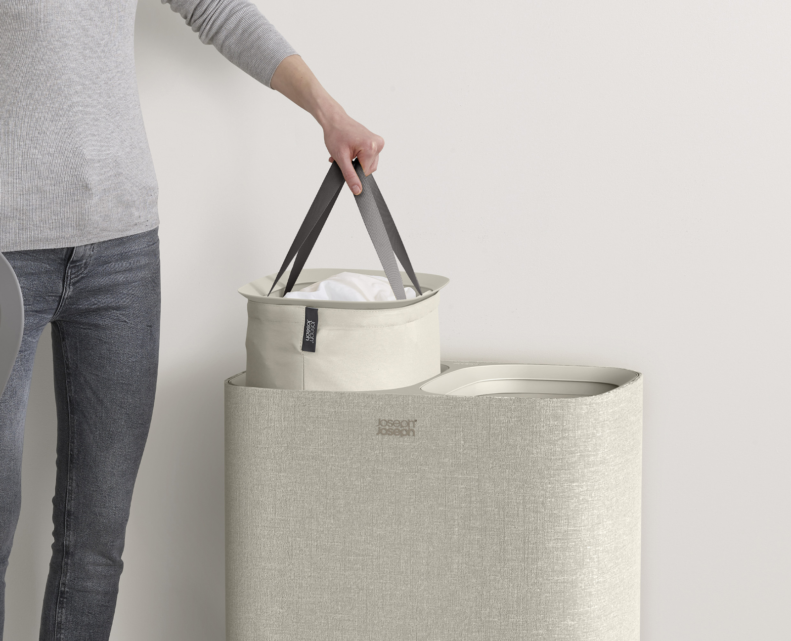 Una persona sostiene una cesta de ropa. La cesta es blanquecina con asas oscuras. El cesto de la ropa es beige.