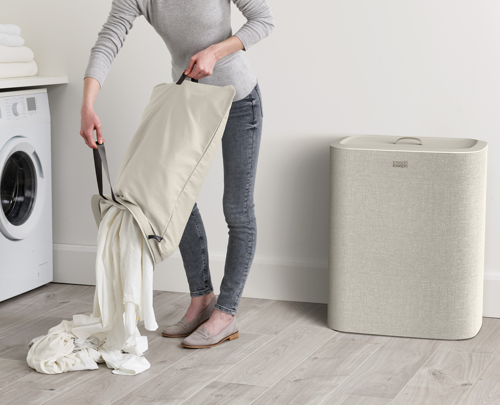 Una persona está vaciando una bolsa de lavandería llena de ropa blanca. Una lavadora y una cesta de ropa sucia están en el fondo.