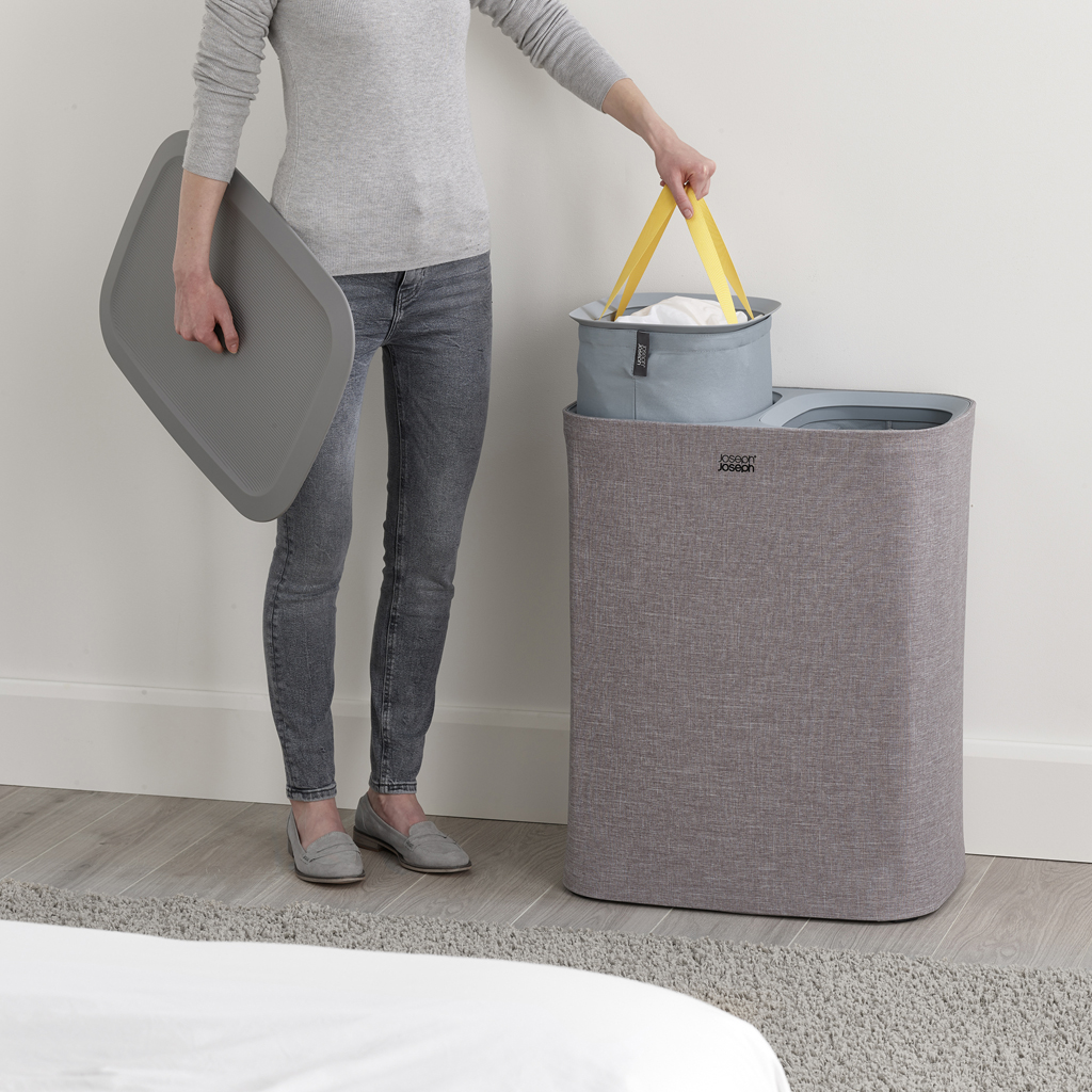 Una persona sostiene una tapa gris y una cesta de lavandería con asas amarillas. Una cesta gris está a su lado, cerca de una pared blanca.
