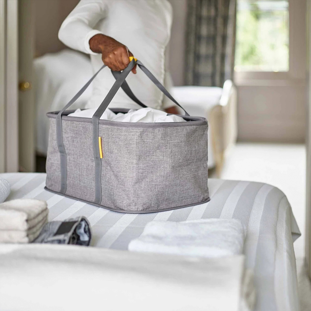 Persona sosteniendo una cesta de lavandería gris llena de sábanas blancas. La cesta está sobre una cama con toallas. El fondo muestra otra cama.