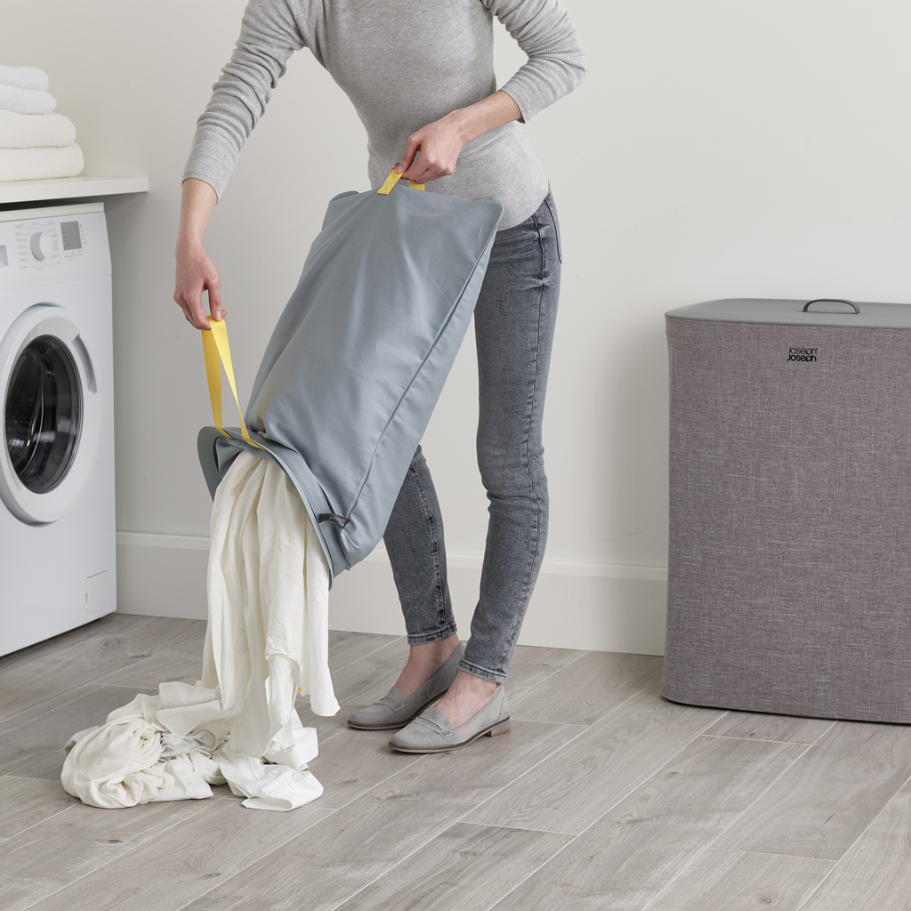 Una persona vacía una bolsa de lavandería gris con ropa blanca. Una lavadora y una cesta de lavandería gris están en el fondo.