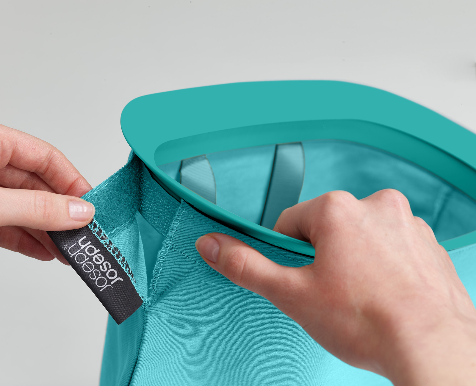 Las manos están ajustando una bolsa Joseph Joseph de color verde azulado. La bolsa es verde azulado y tiene una etiqueta oscura con el nombre de la marca.