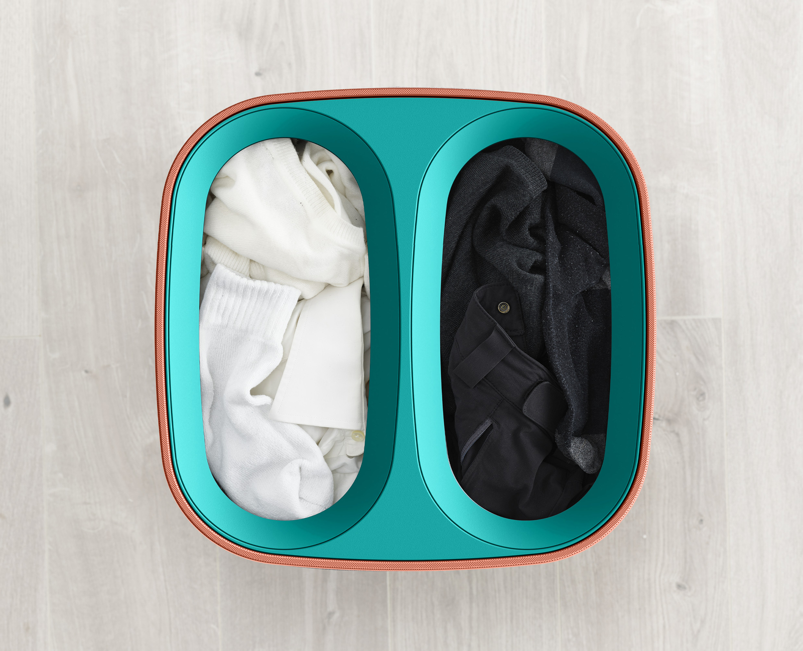 Una cesta de lavandería dividida contiene ropa blanca y negra. La cesta es de color verde azulado con un borde naranja, sobre un piso de madera.