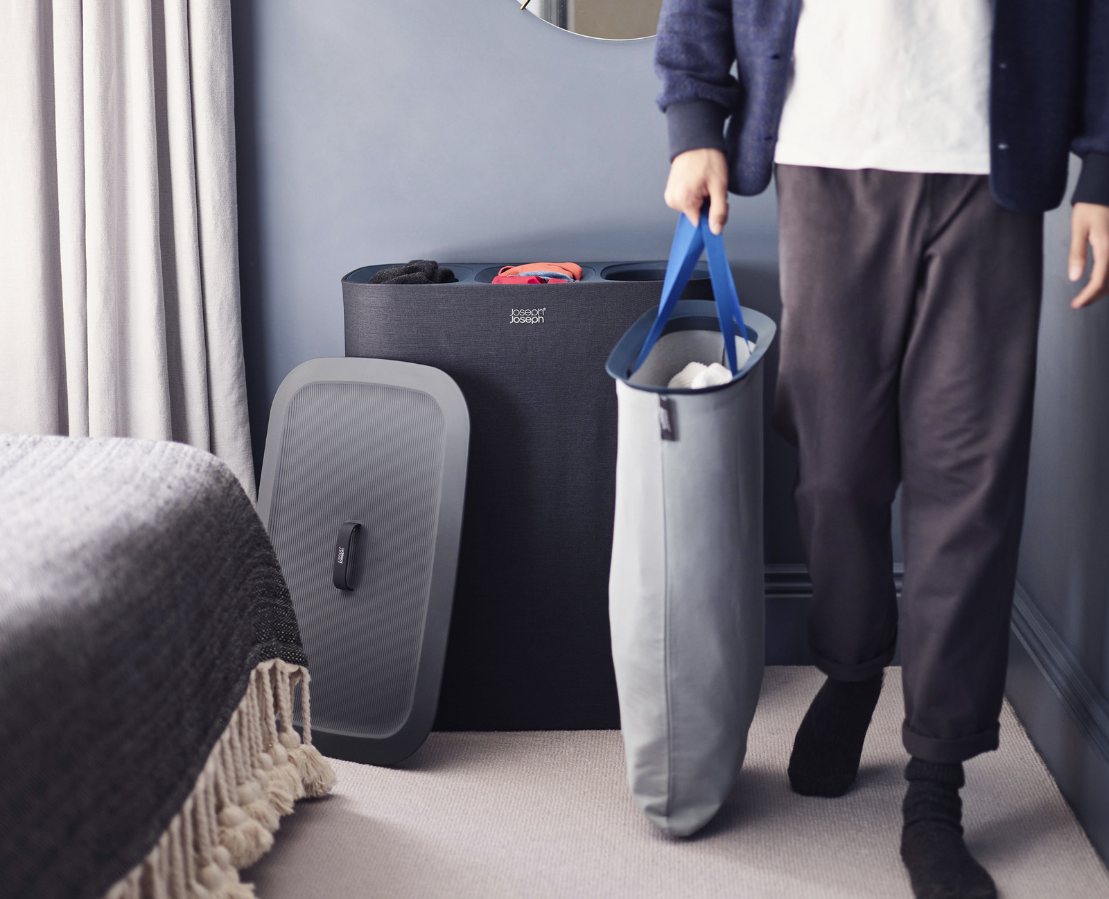 Una persona lleva una bolsa de lavandería. Un clasificador de ropa sucia y una tapa están cerca. La escena es interior, con cama y cortinas.