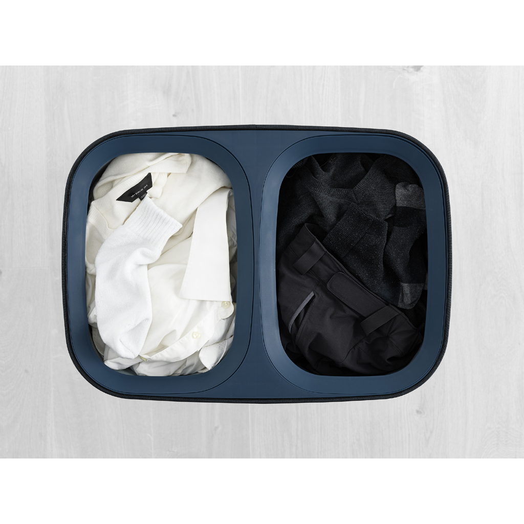 Una cesta de lavandería dividida llena de camisas blancas, un calcetín y ropa negra. La cesta es azul, sobre un piso de madera clara.