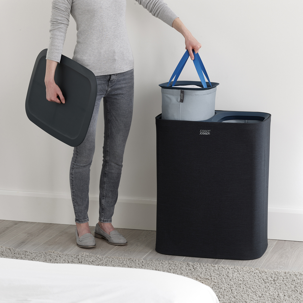 Una persona sostiene una tapa gris oscura y una cesta de lavandería con asa azul. Una cesta de lavandería negra está a su lado, en una habitación luminosa.