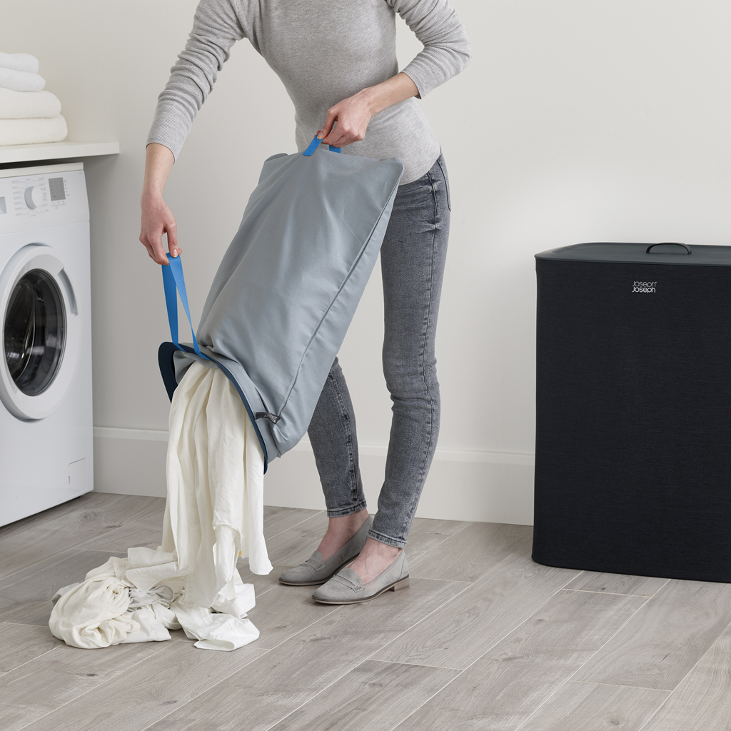 Una persona está vaciando ropa de una bolsa de lavandería gris. También se ve una lavadora y una cesta de ropa negra.